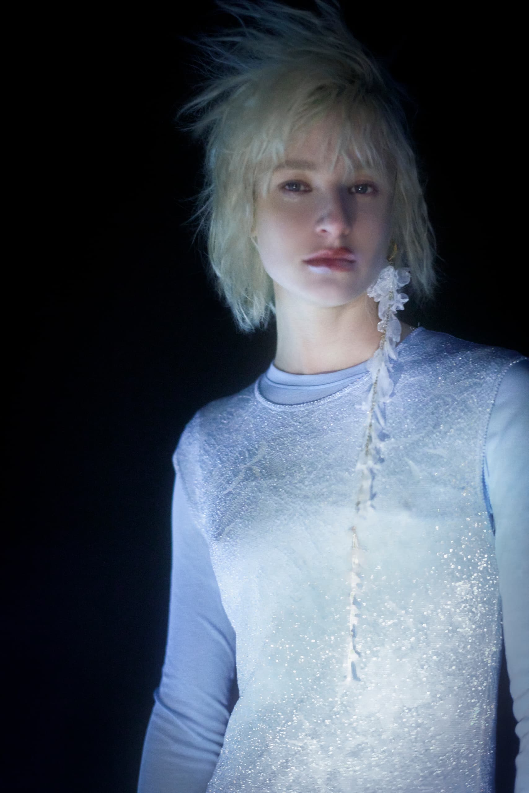 Glamorous person in shimmering outfit, blonde hair lit against a dark background.