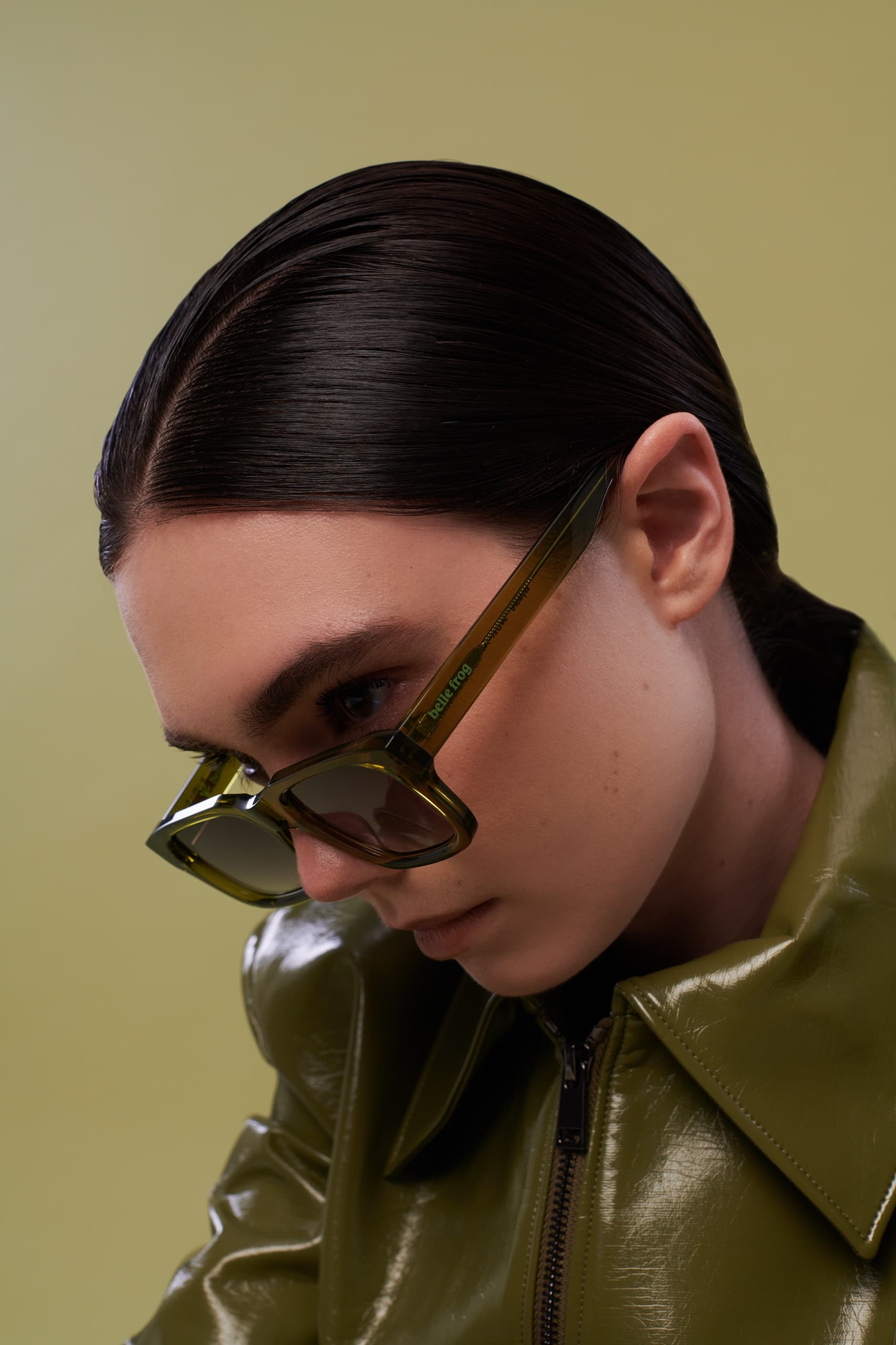 Woman in green sunglasses and jacket, sleek hairstyle, neutral background.