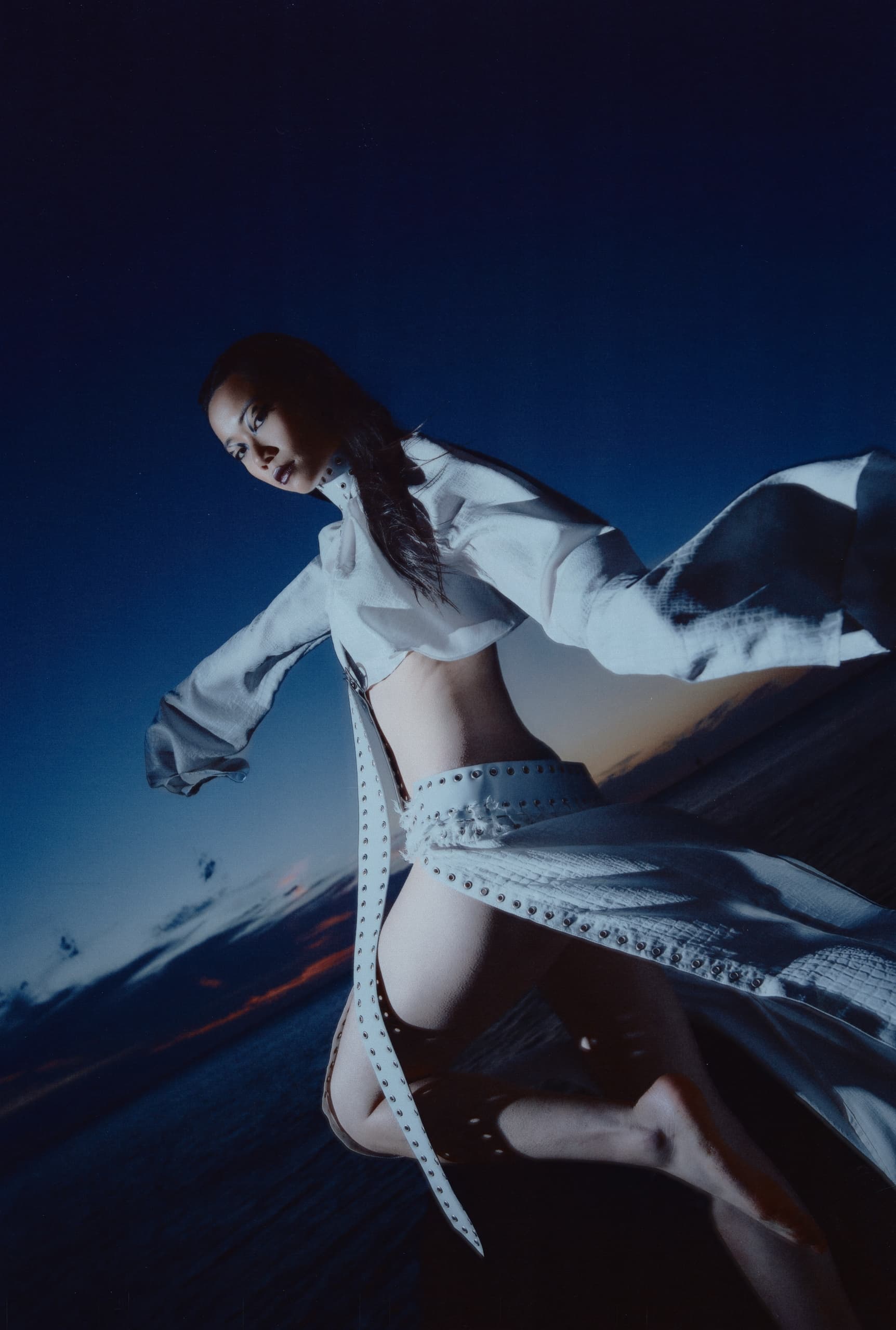 Fashion model in dynamic white outfit against a dramatic twilight sky.