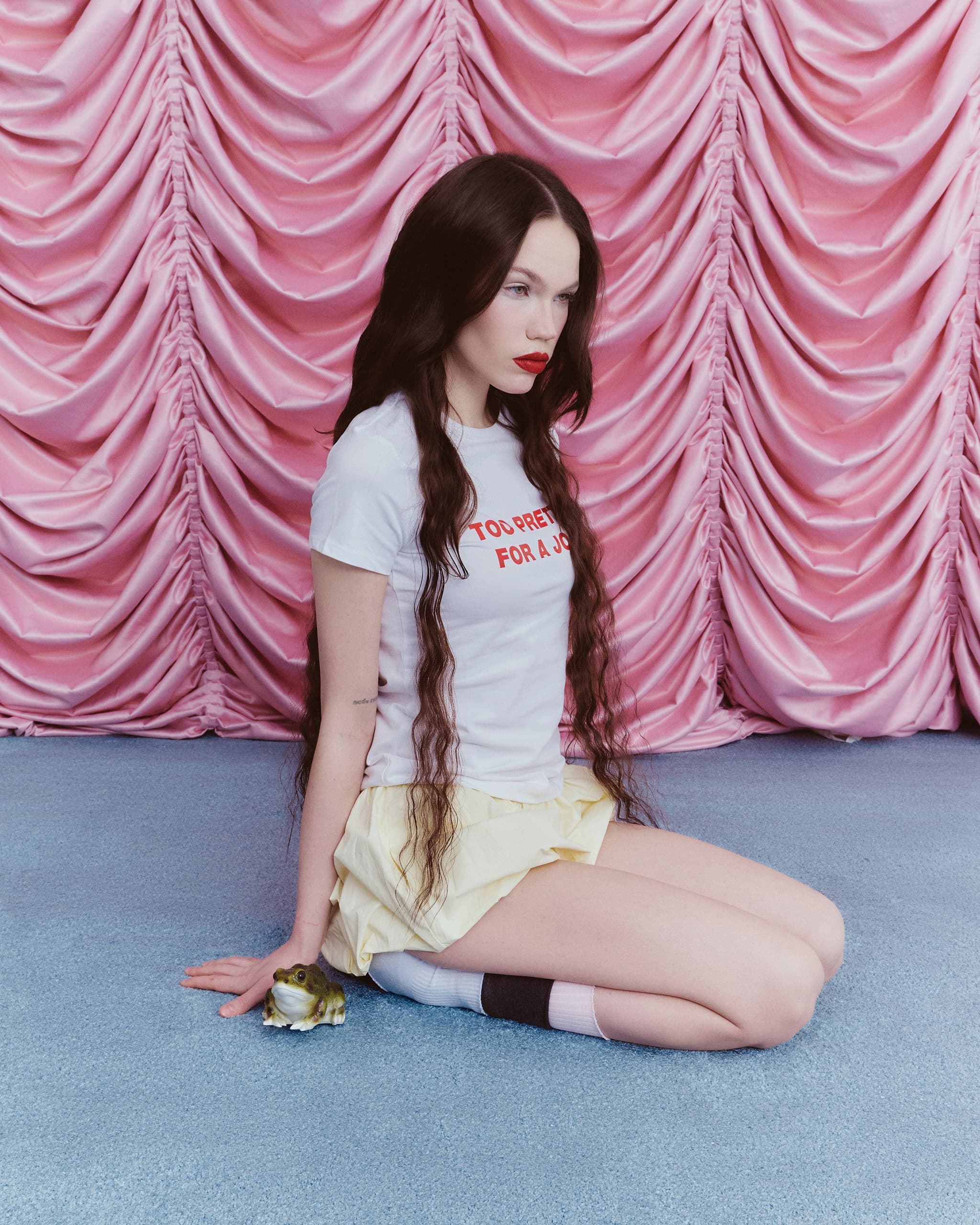 Person sits on blue floor in front of pink curtain, next to a frog statue.