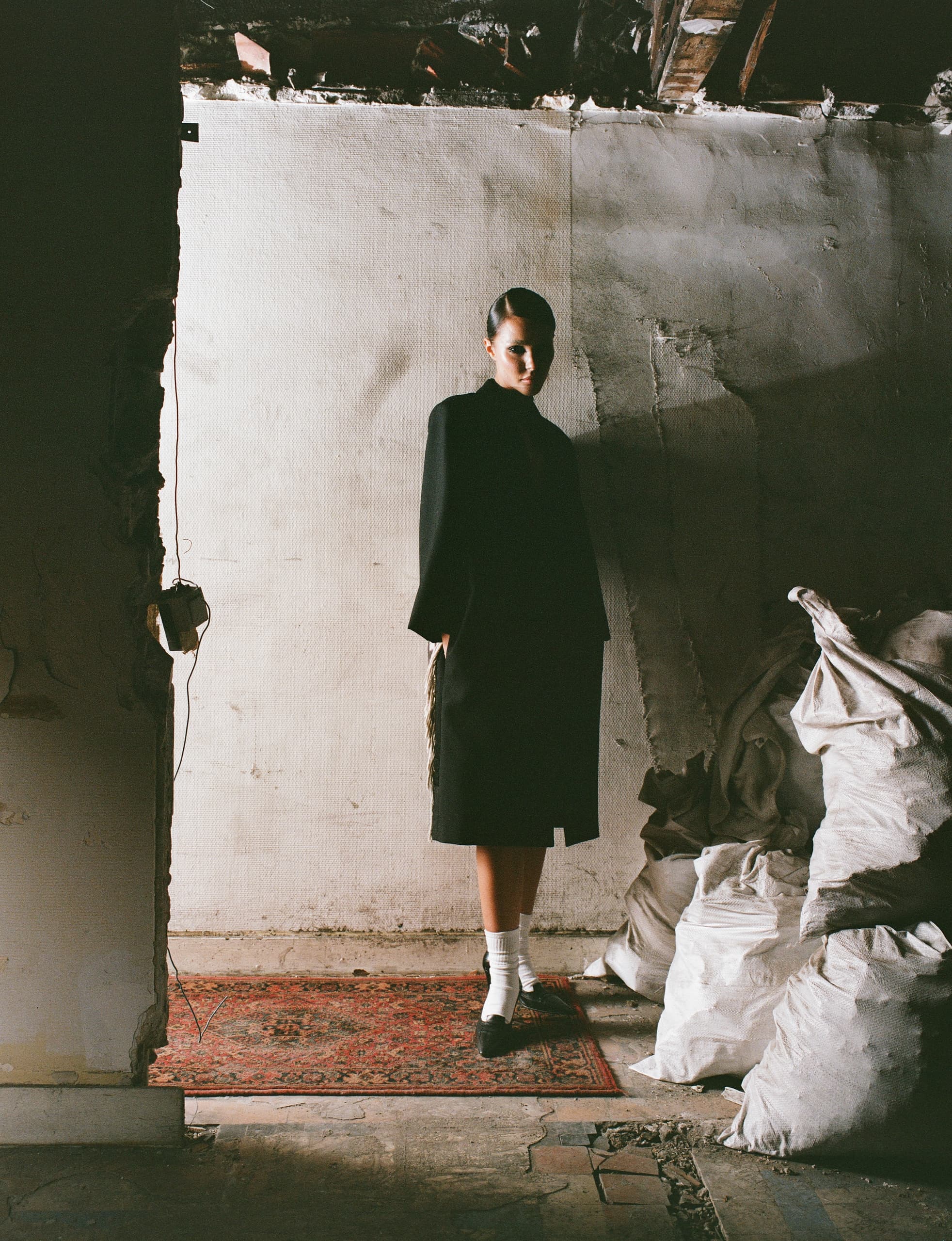 Woman in dark coat stands on vintage rug in rustic, moody setting.