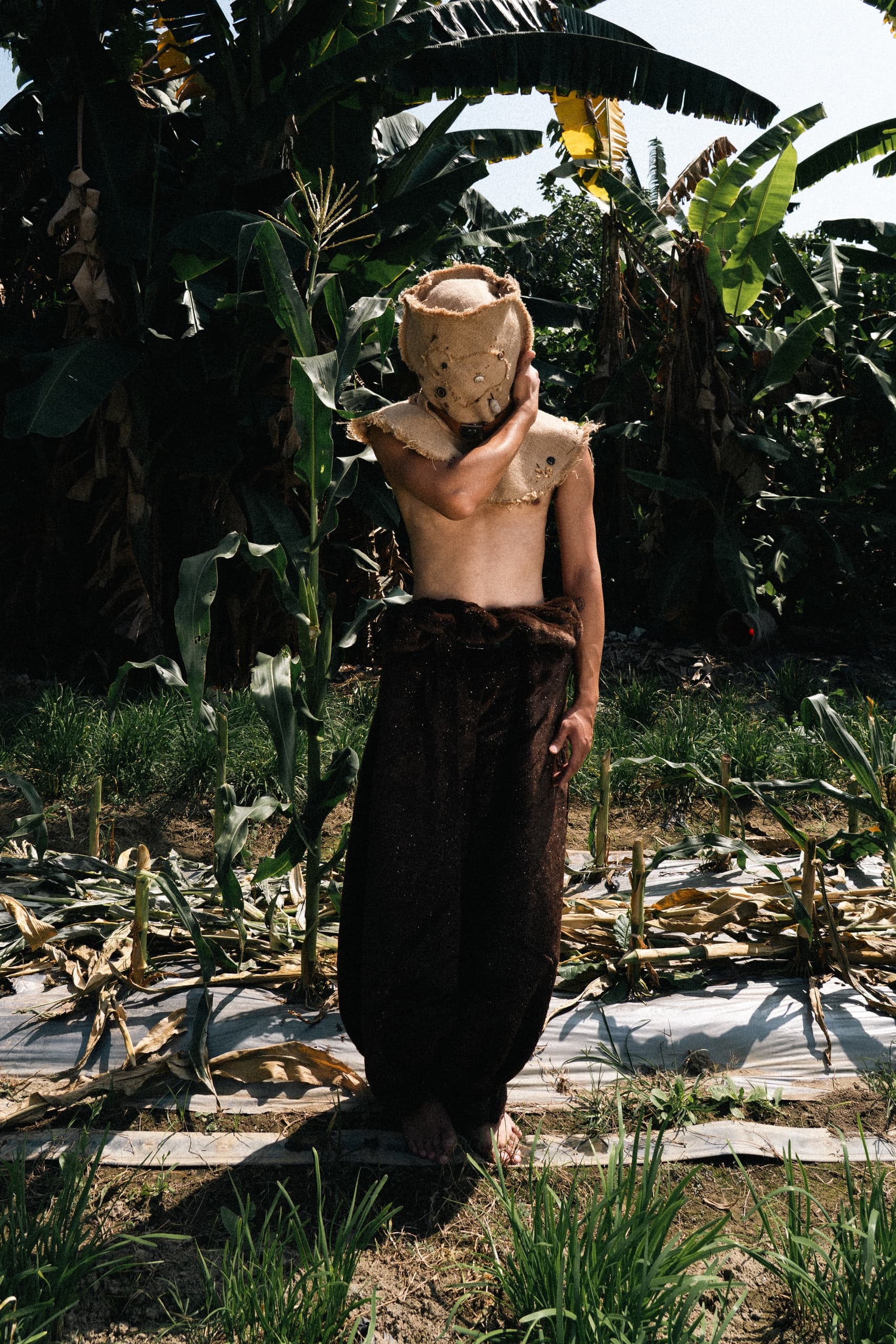 Person wearing burlap mask in tropical garden, surrounded by banana plants.