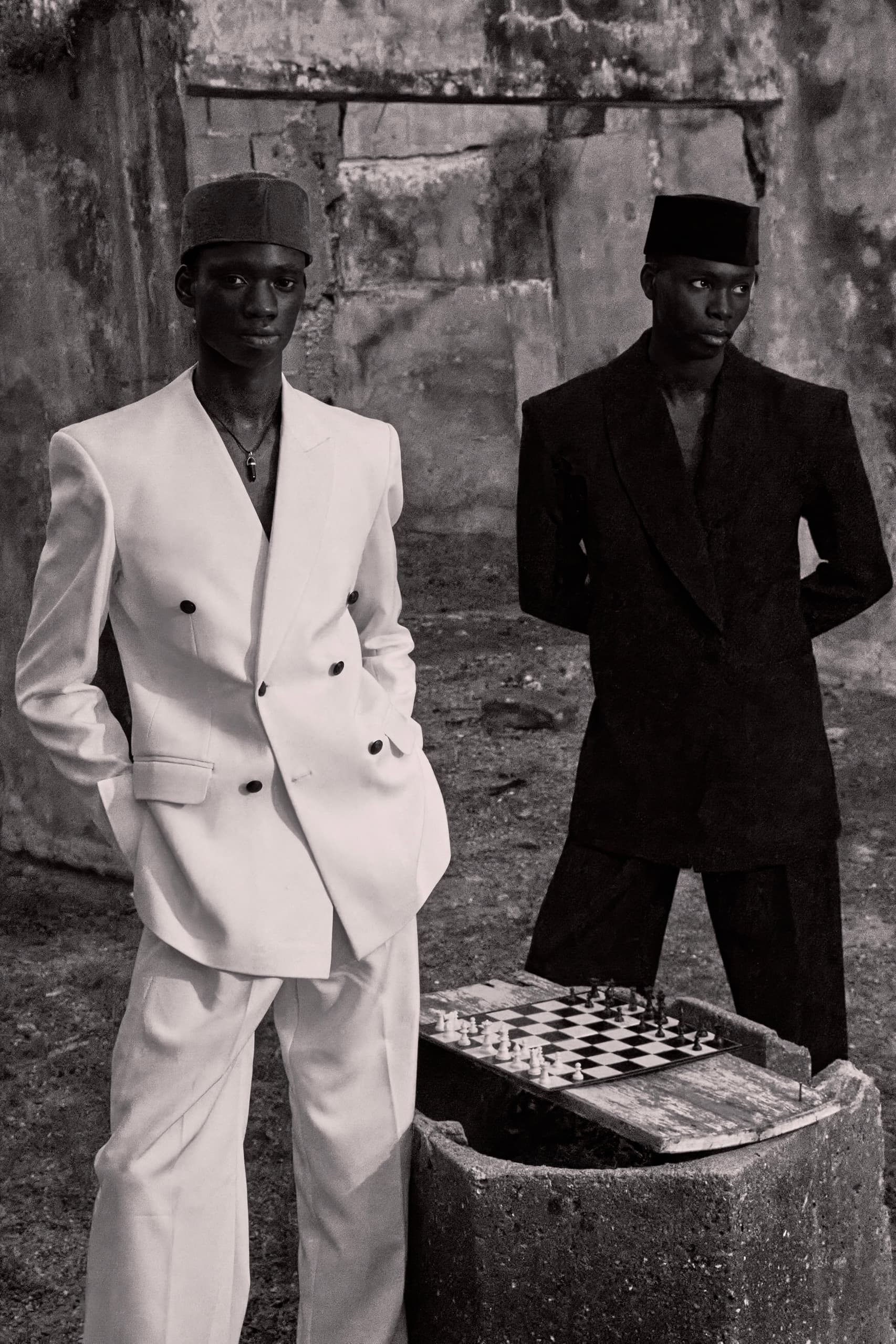 Stylish men in suits beside rustic chessboard outdoors, black and white photography.