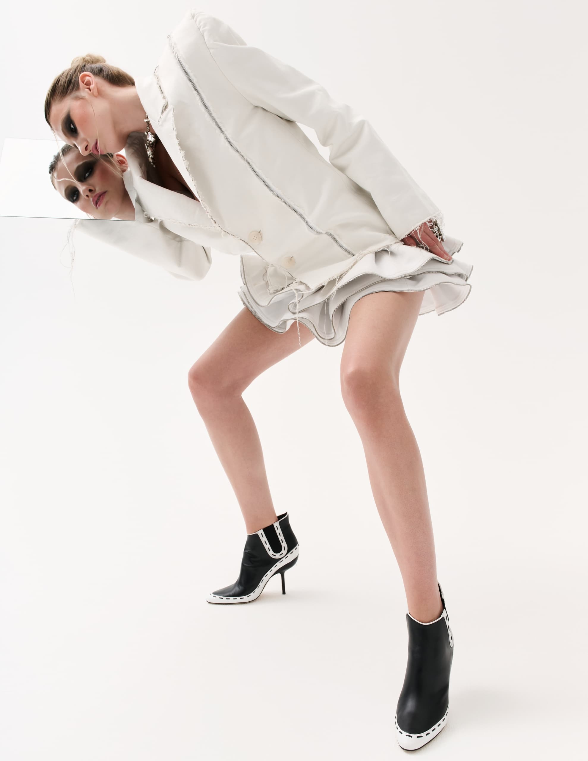 Fashionable woman in white blazer and black boots poses with mirror.