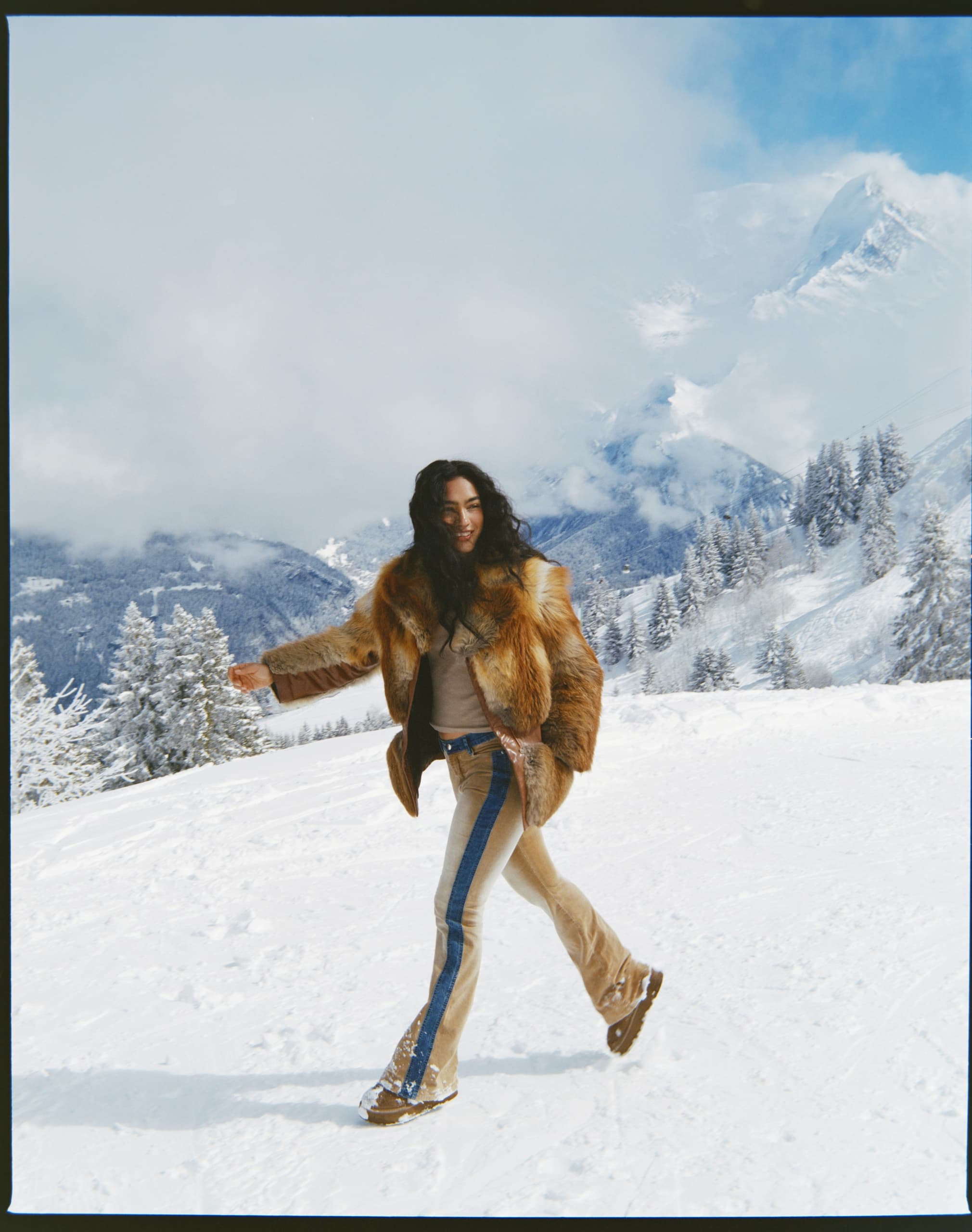 Person in fur coat walks joyfully through snowy mountain landscape.