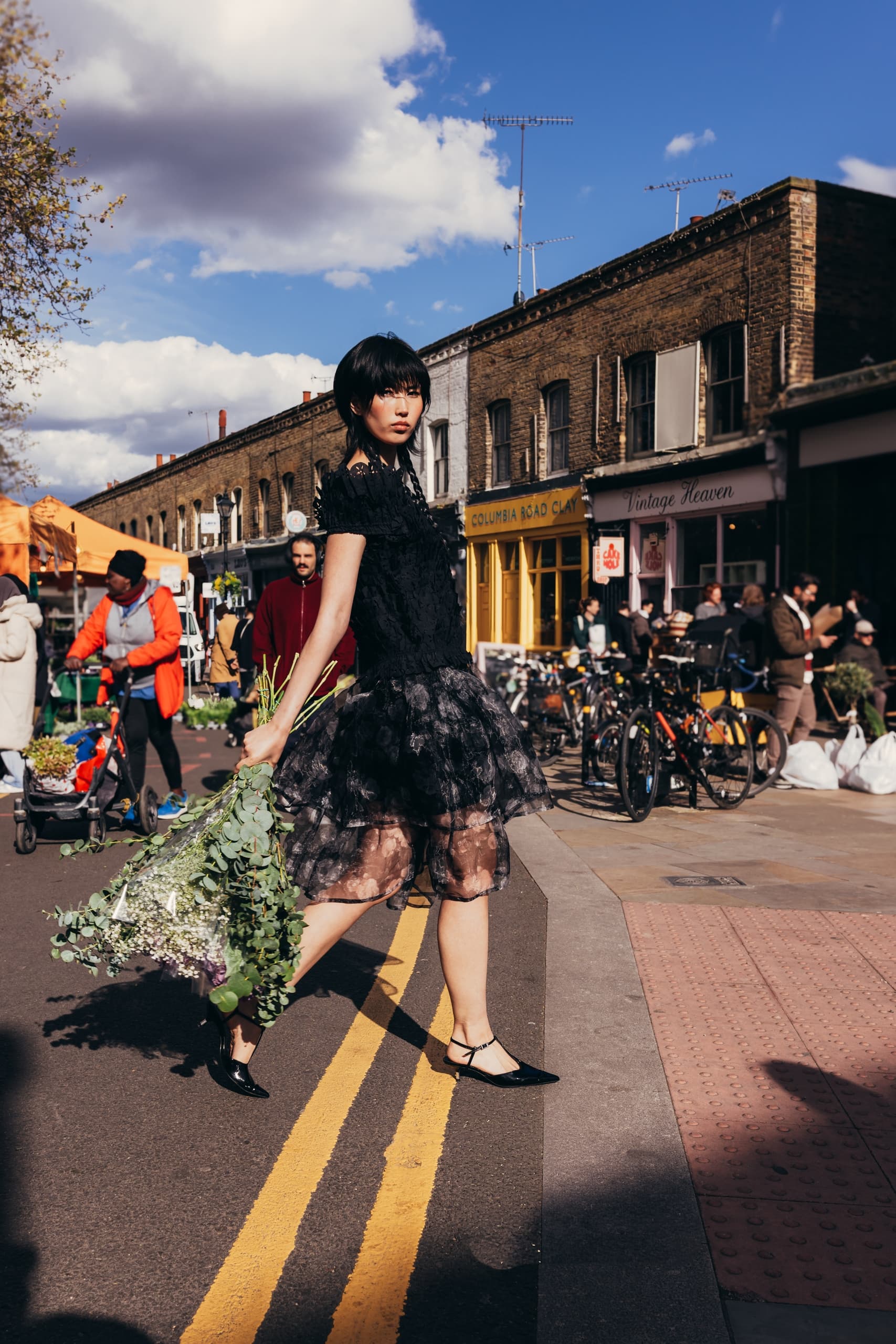 Fashionable woman with bouquet walking through vibrant city street market.
