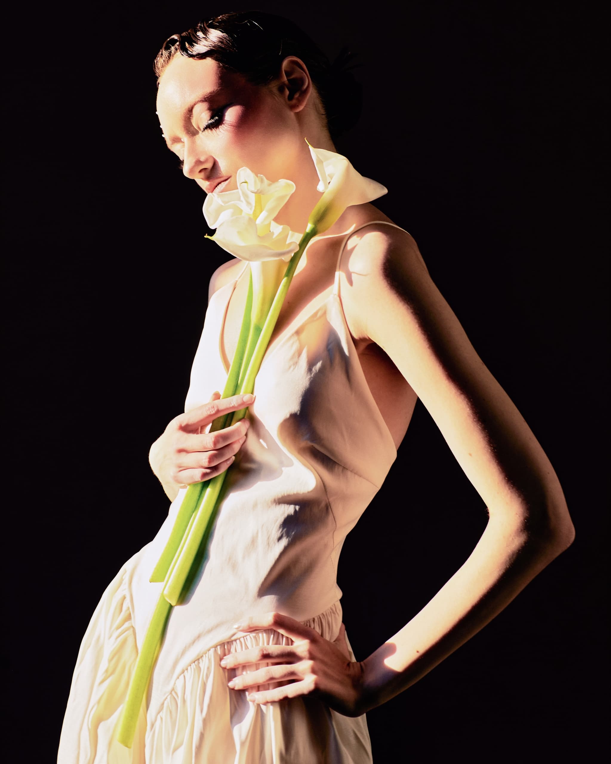 Woman in soft lighting holding white calla lilies against a dark background.
