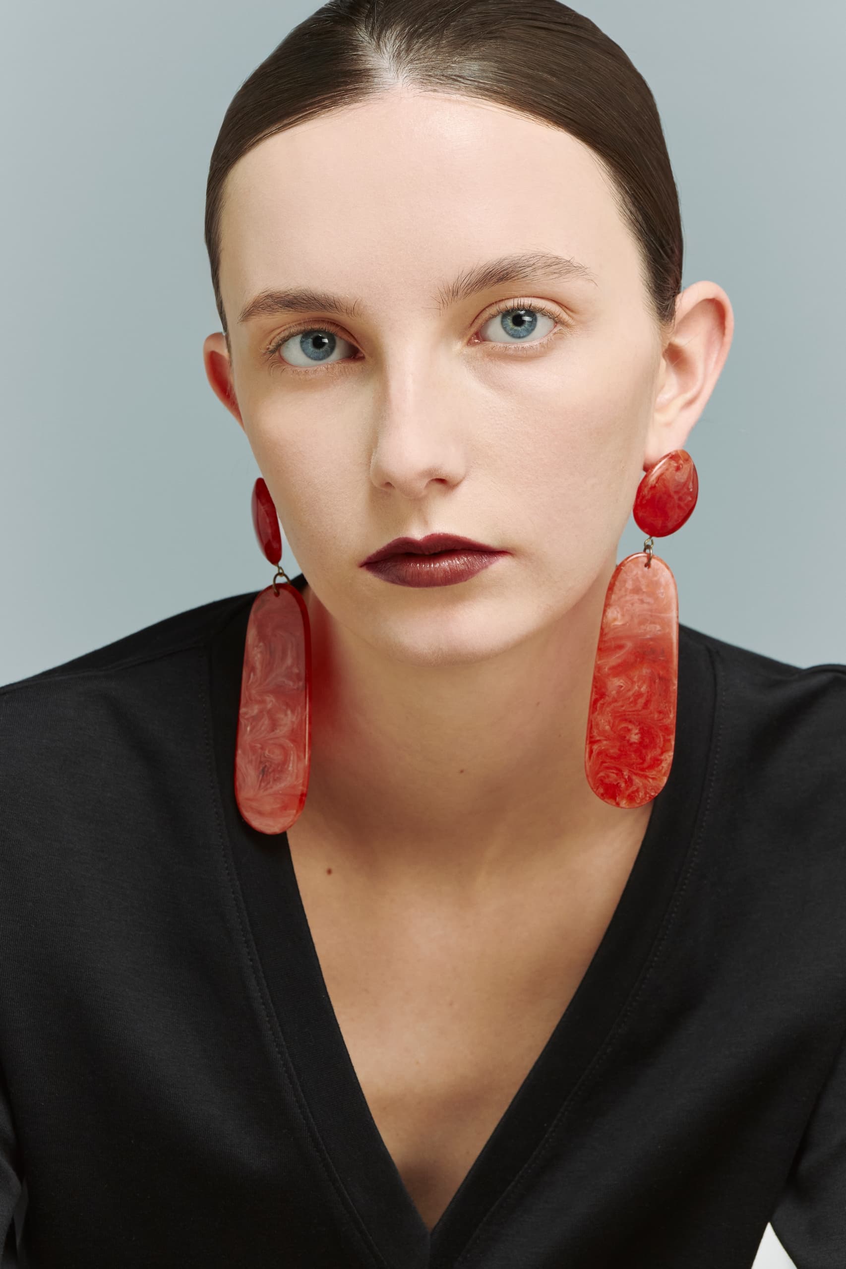Woman wearing large red earrings and dark top, with sleek hair and neutral backdrop.