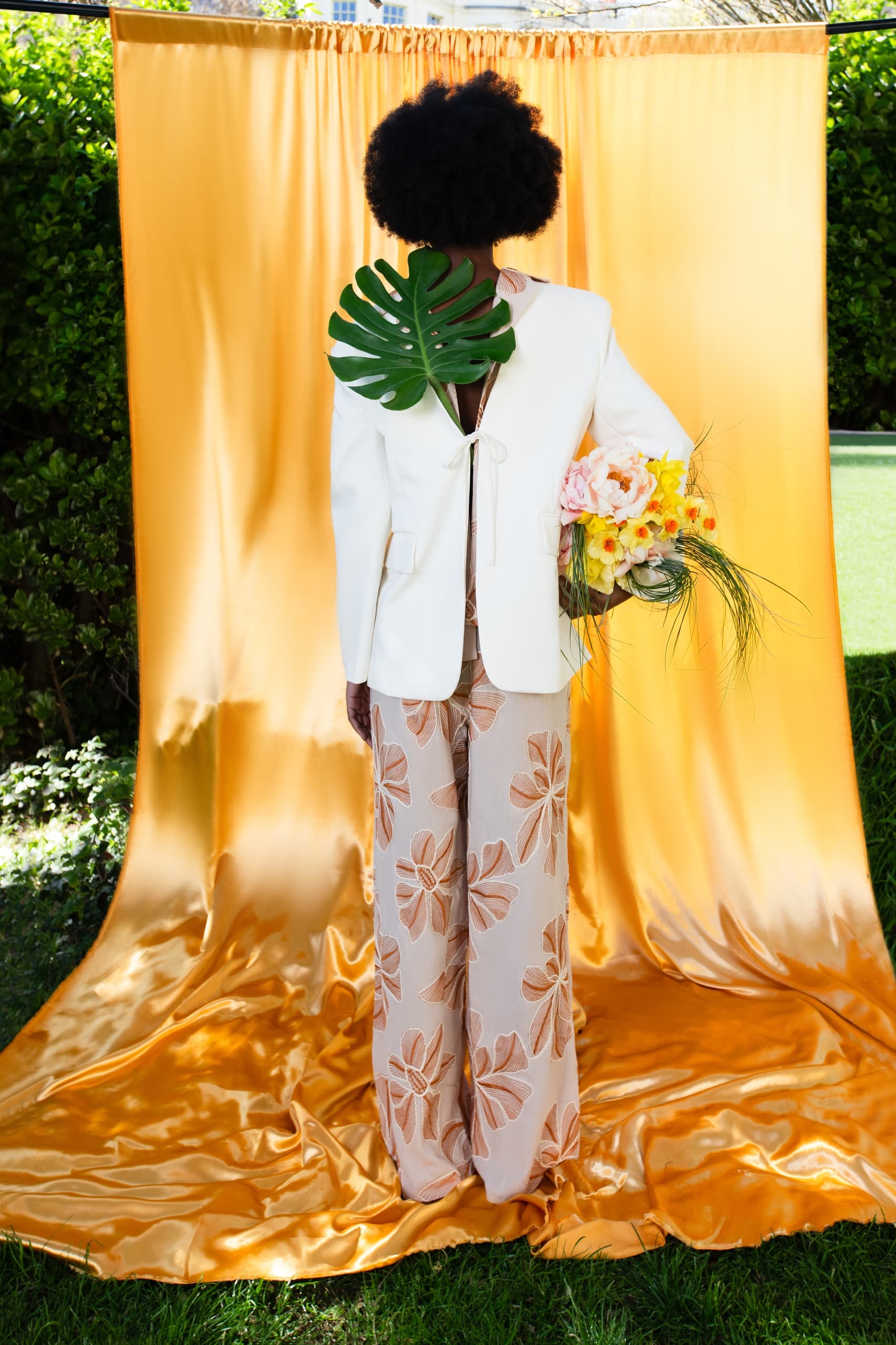 Fashion model in floral outfit holding bouquet against gold backdrop.
