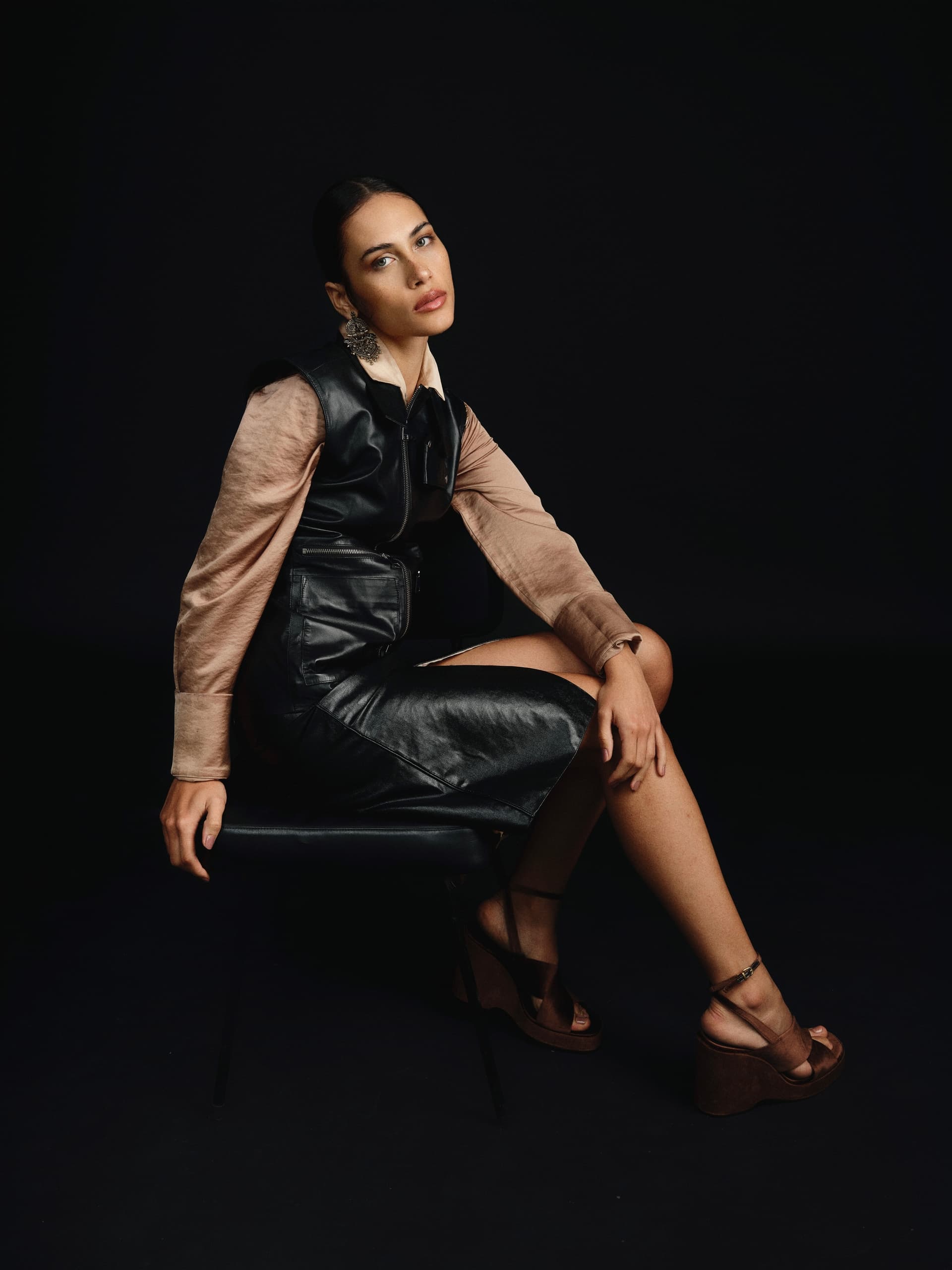 Elegant woman in black leather dress poses stylishly on a dark chair.