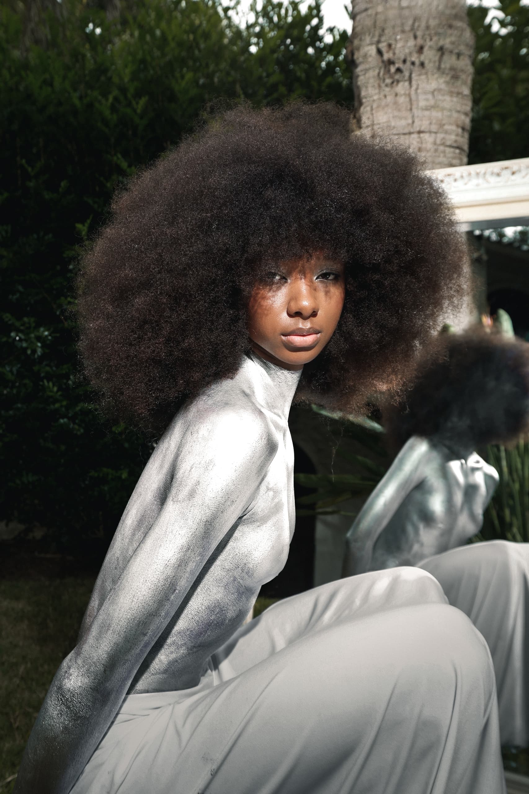 Model with silver body paint and afro hair poses outdoors, mirror reflection visible.