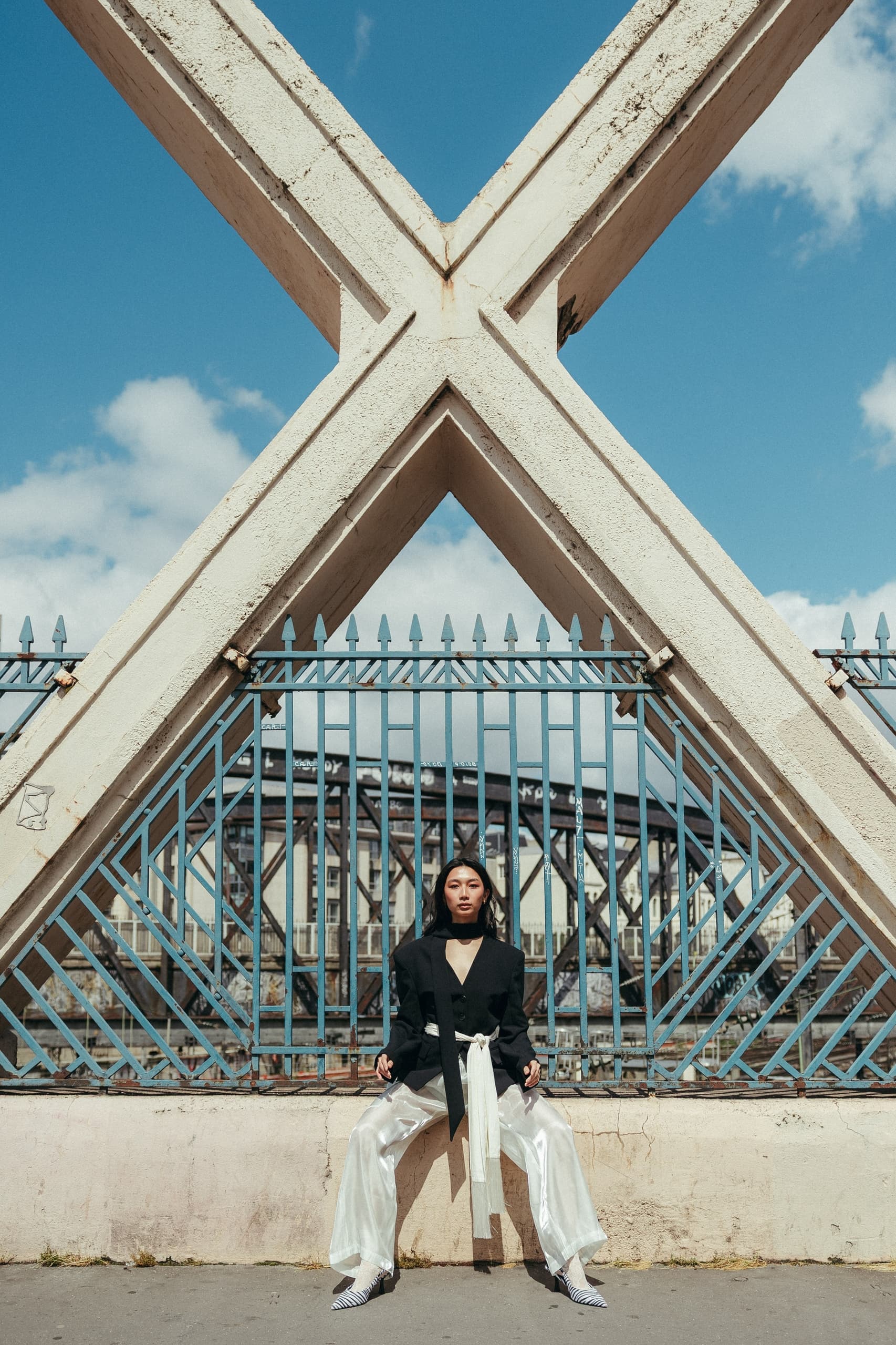 Fashionable woman in chic outfit sitting under geometric structure, urban backdrop.