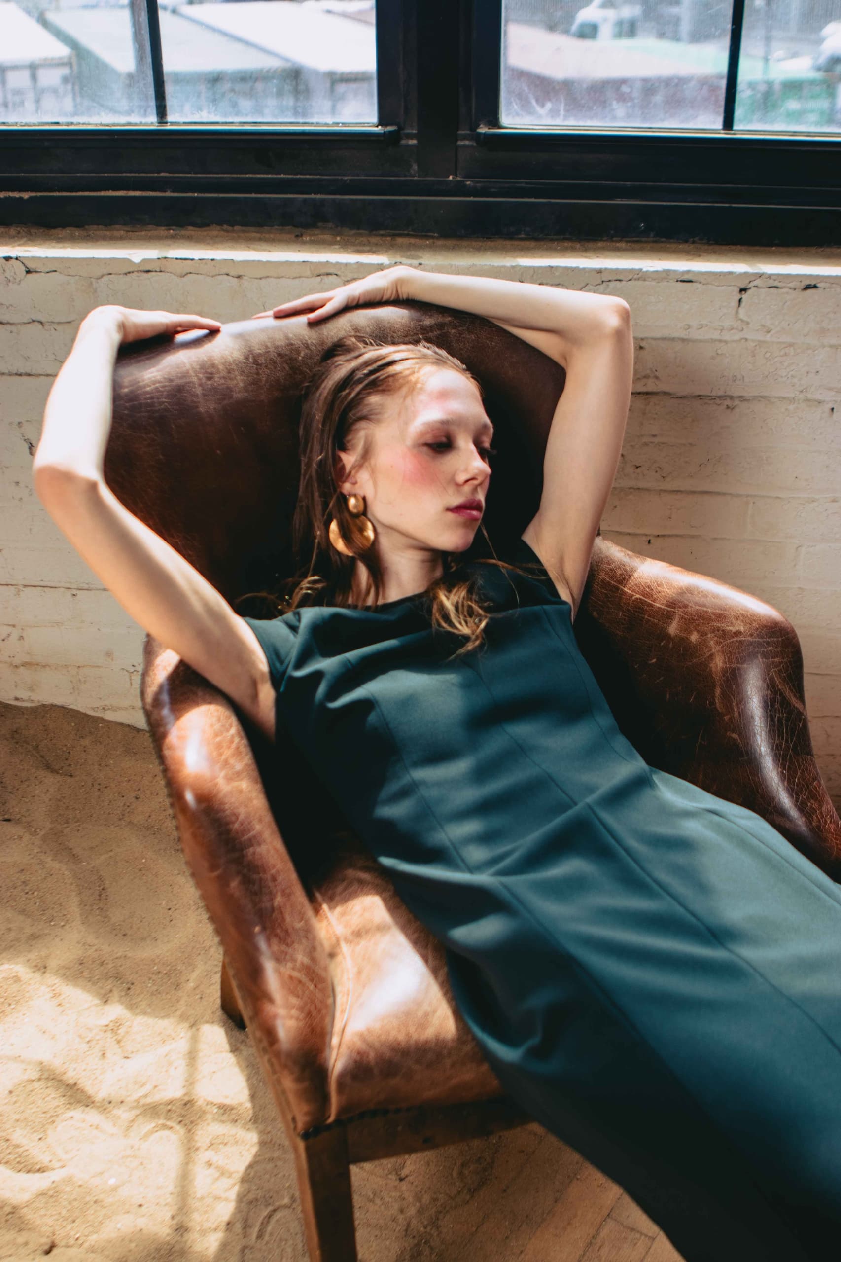 Woman relaxing in an armchair by sunlit window, wearing a dark green dress.