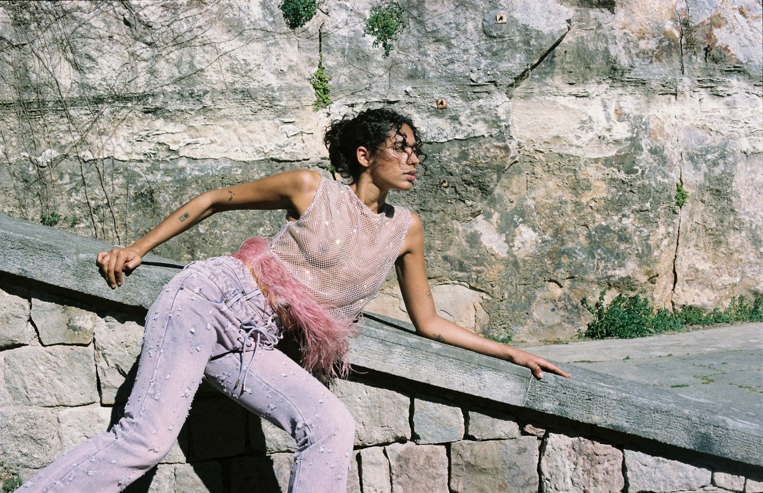 Stylish person in pink feathered outfit leaning on stone wall.