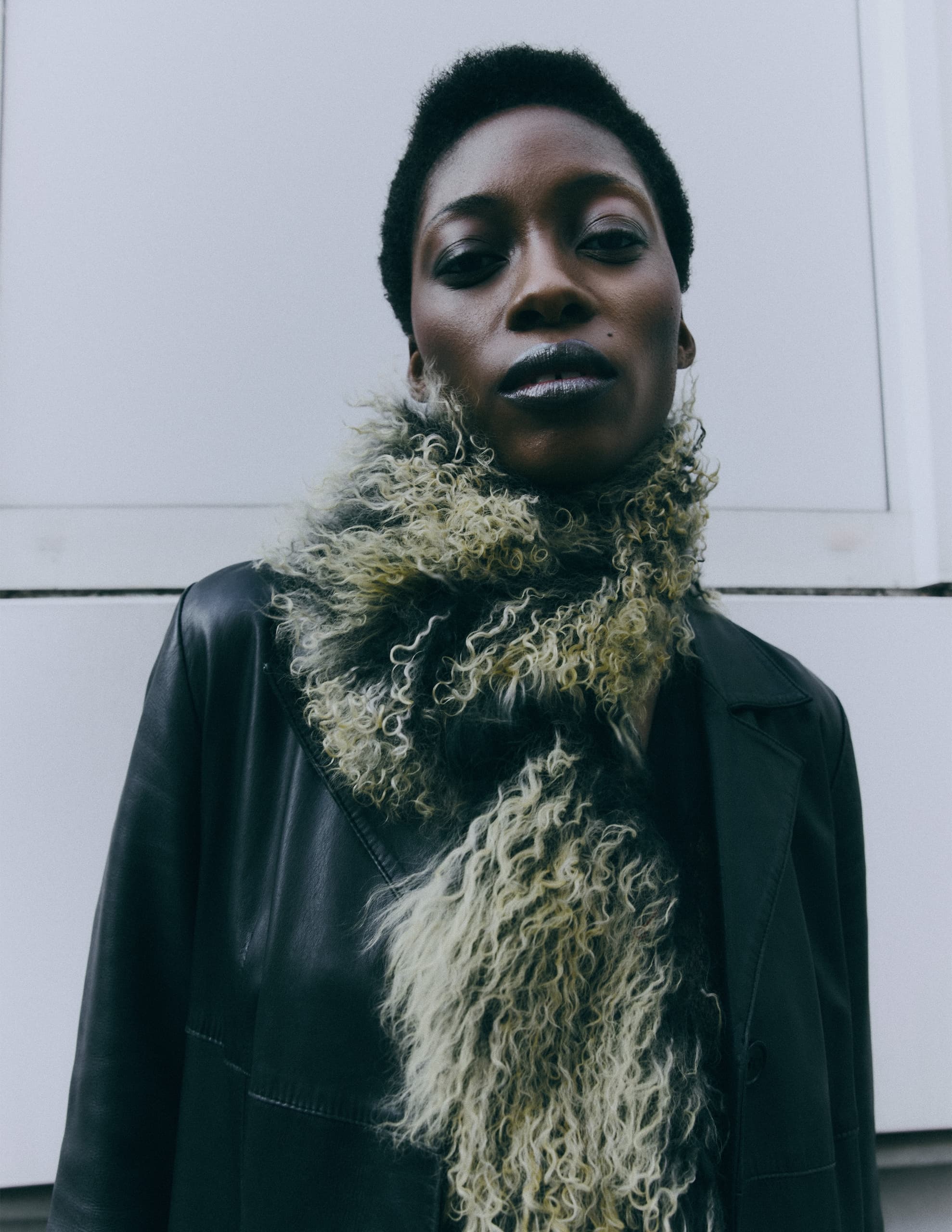 Fashionable person in leather jacket and curly scarf, standing against a white backdrop.