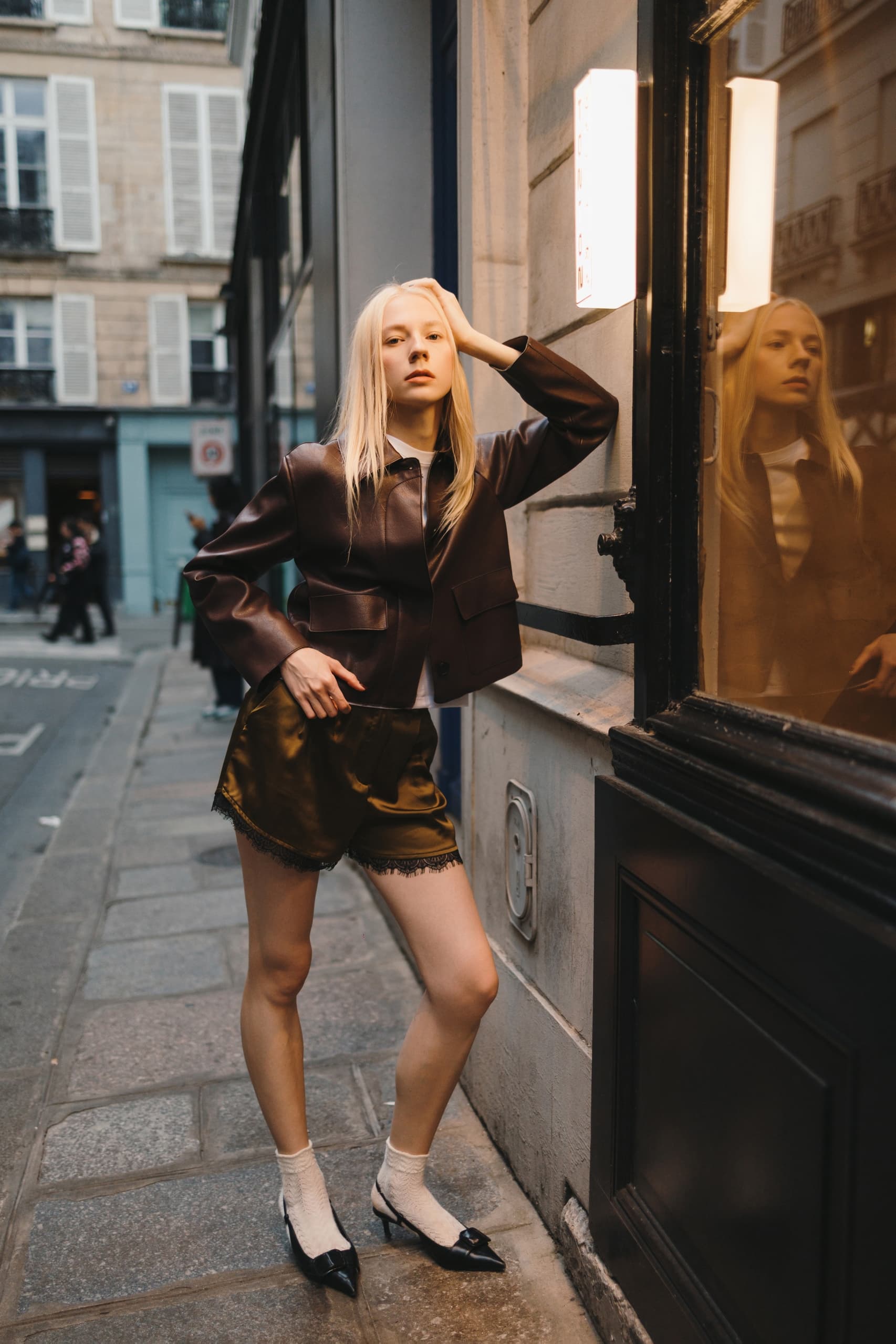 Stylish woman in leather jacket posing by a city street window at dusk.