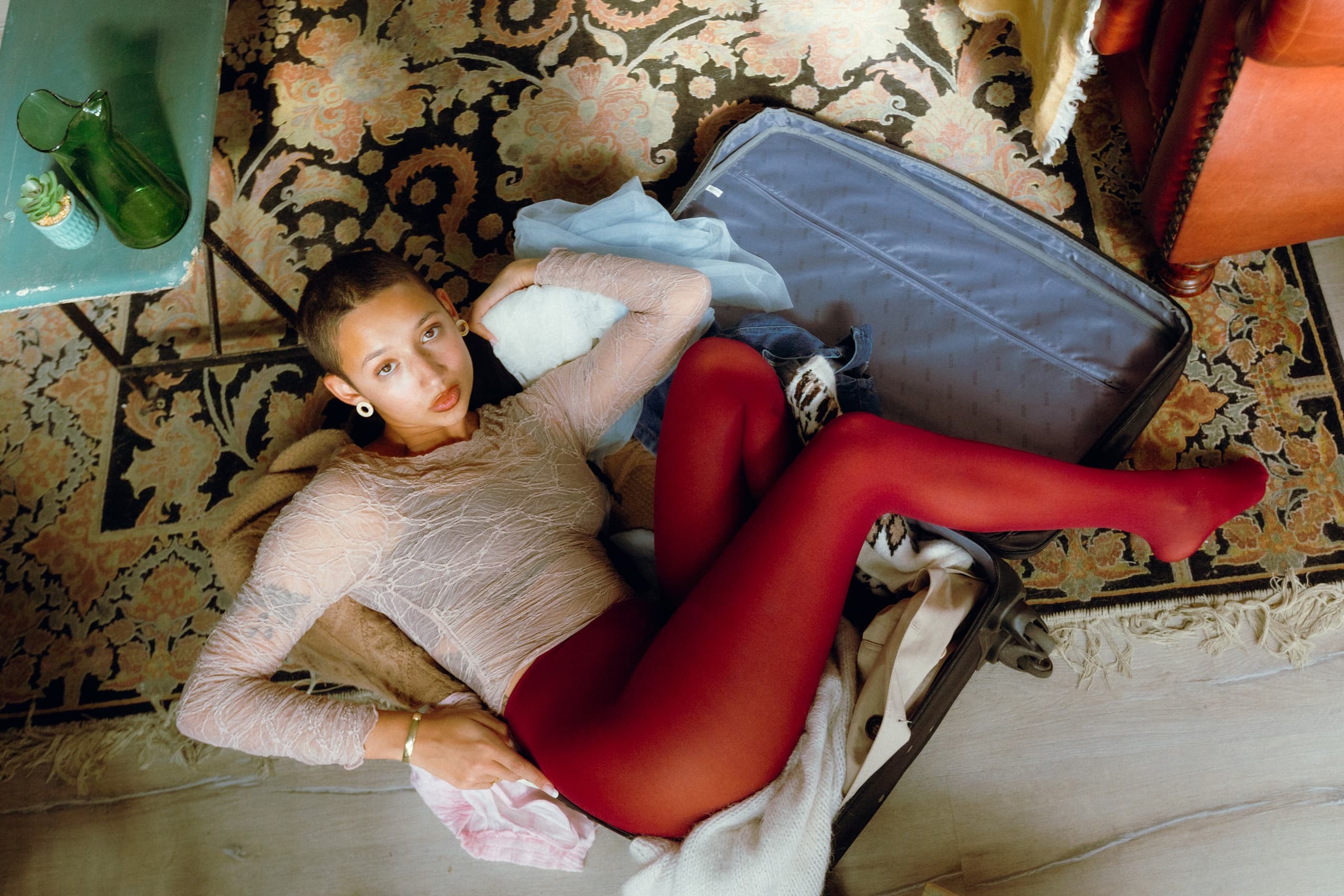 Person lounging in an open suitcase on a patterned rug, wearing red tights.