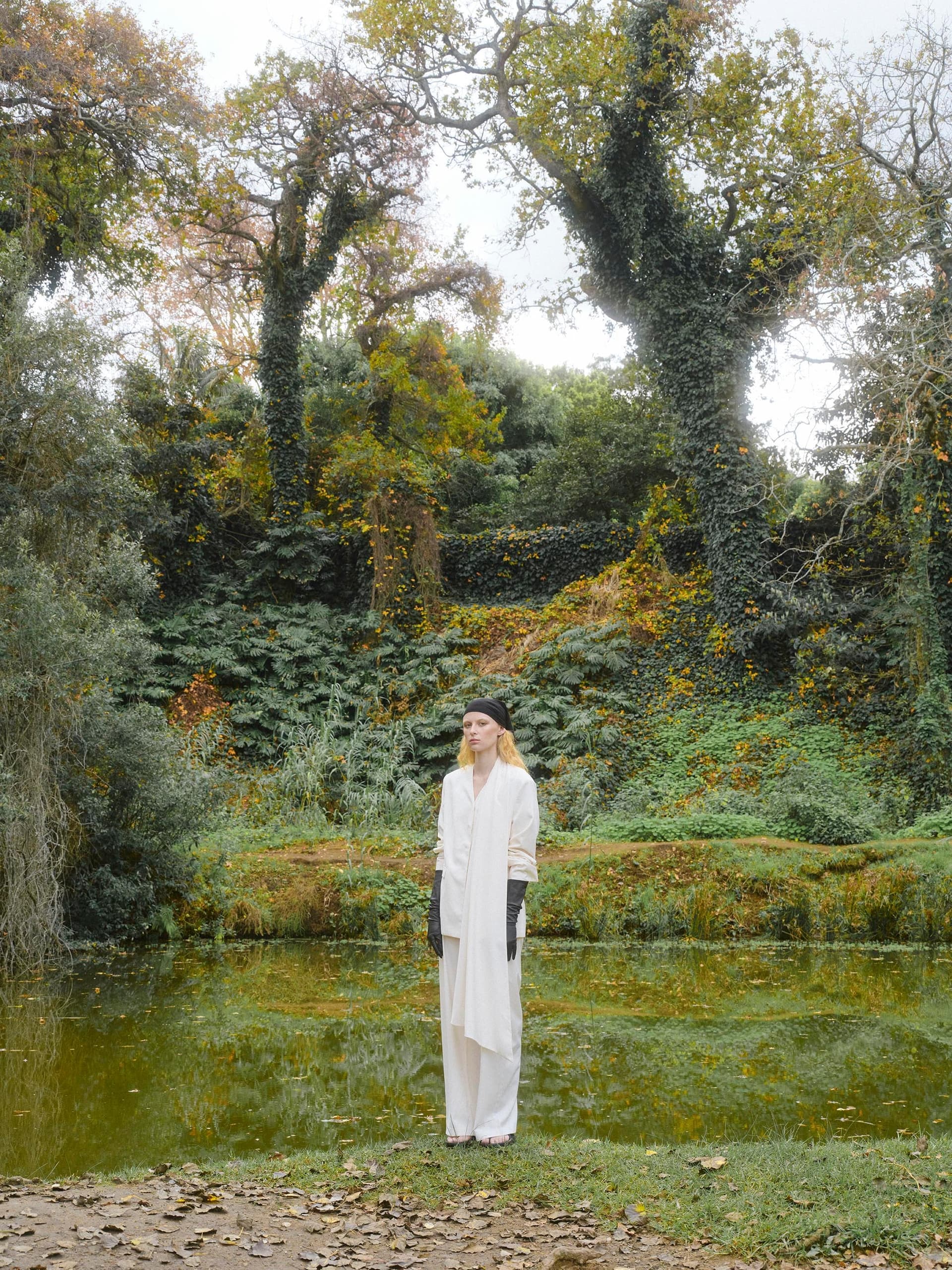 Person in white outfit beside a pond in a lush, leafy forest setting.