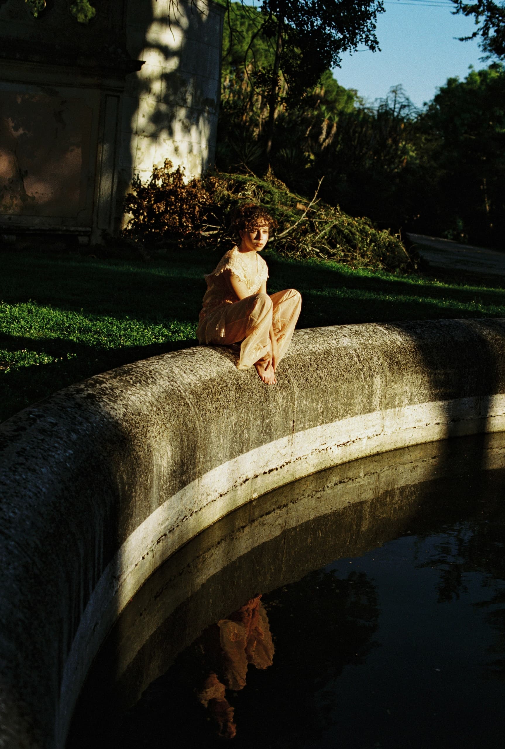 Woman in flowing dress sits by fountain edge, reflecting in still water.
