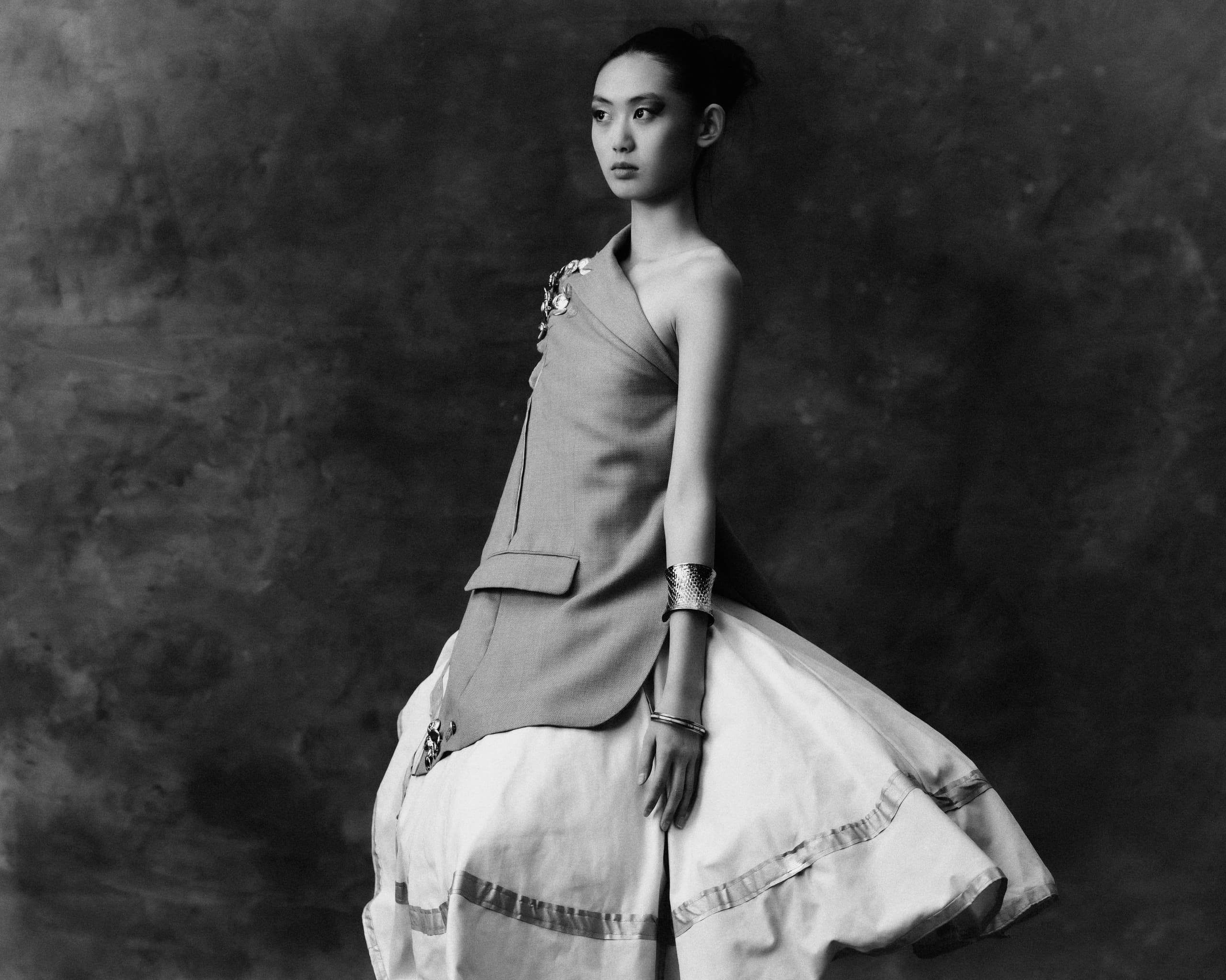 Elegant fashion model in asymmetrical gown poses against a textured background.