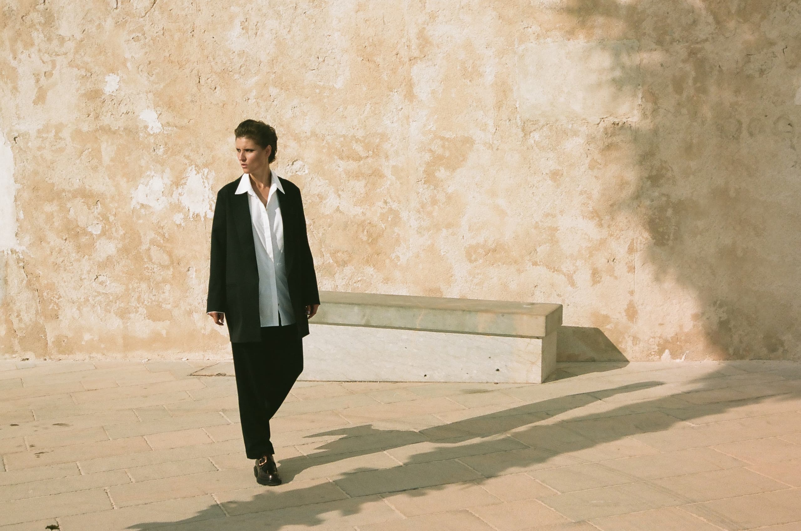 Person in formal attire walking by sunlit textured wall.