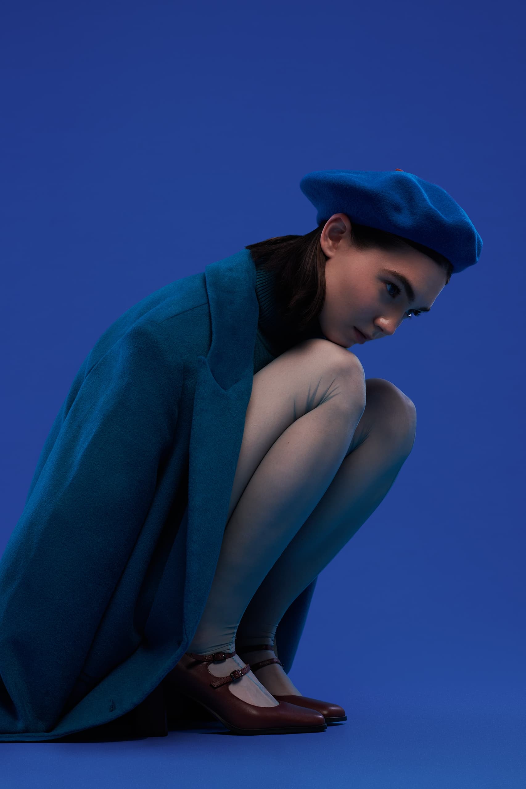 Woman in blue beret and coat crouching, deep in thought against a blue background.