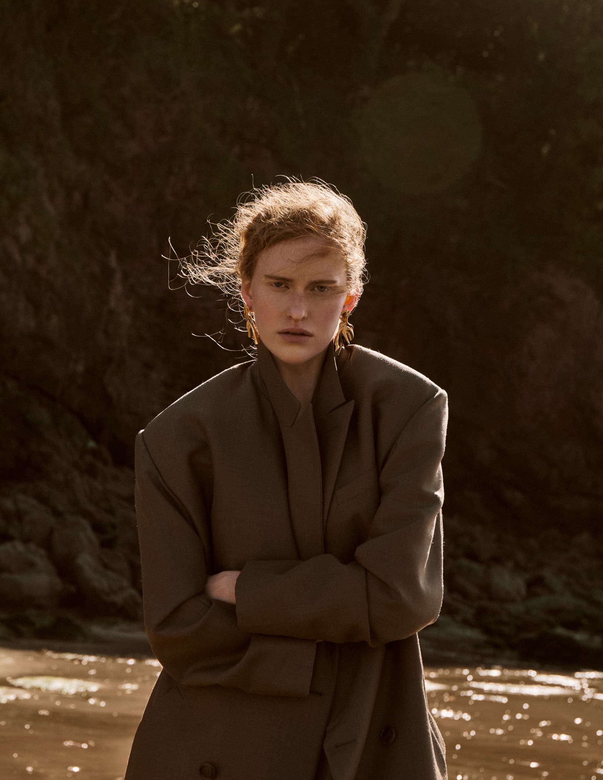 Person in oversized blazer by rocky coast, arms crossed, wind blowing.