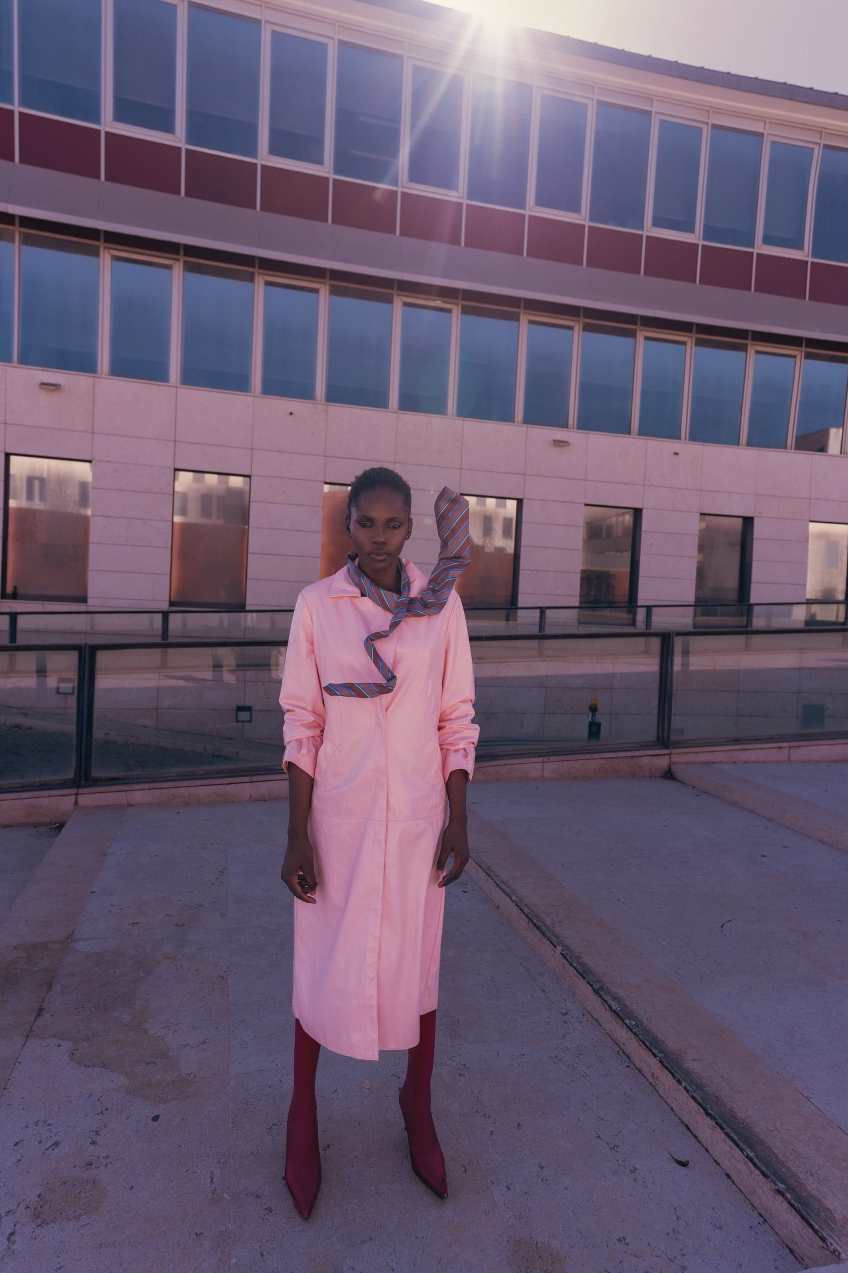 Stylish woman in pink dress and scarf under sunlight near a modern building.