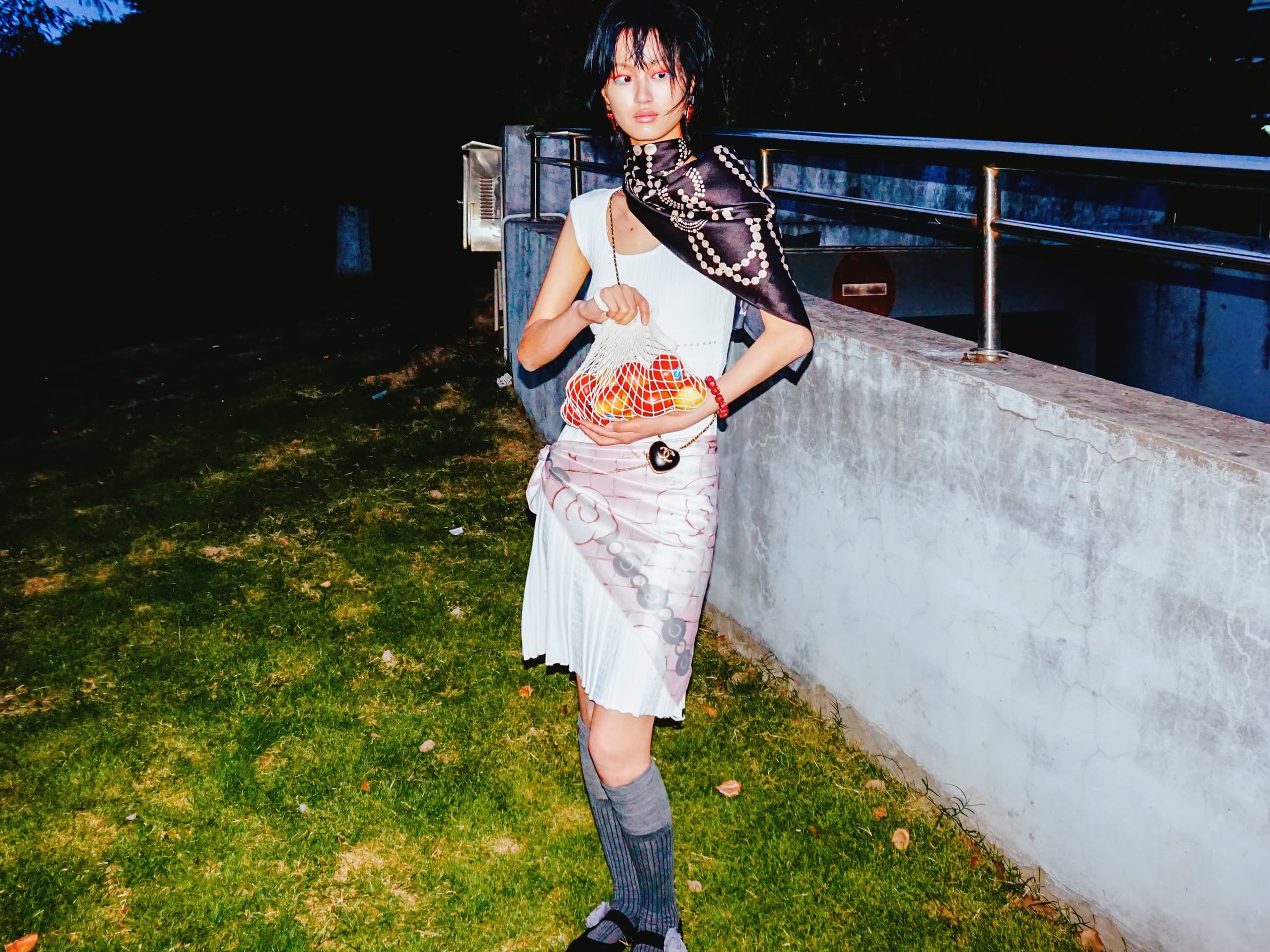 Fashionable woman in eclectic outfit, standing outdoors at night, holding a vibrant handbag.
