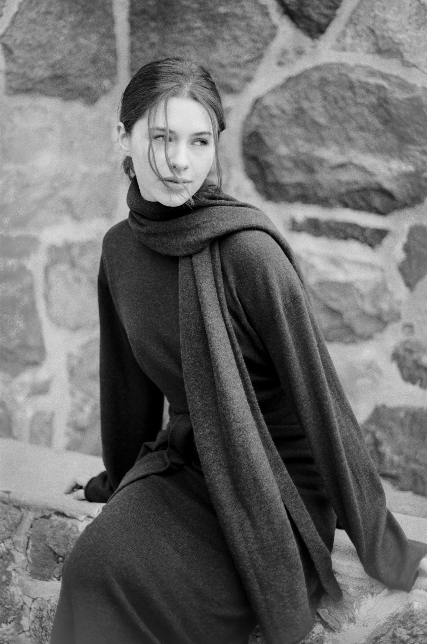 Black and white portrait of a woman in a long coat, sitting by a stone wall.