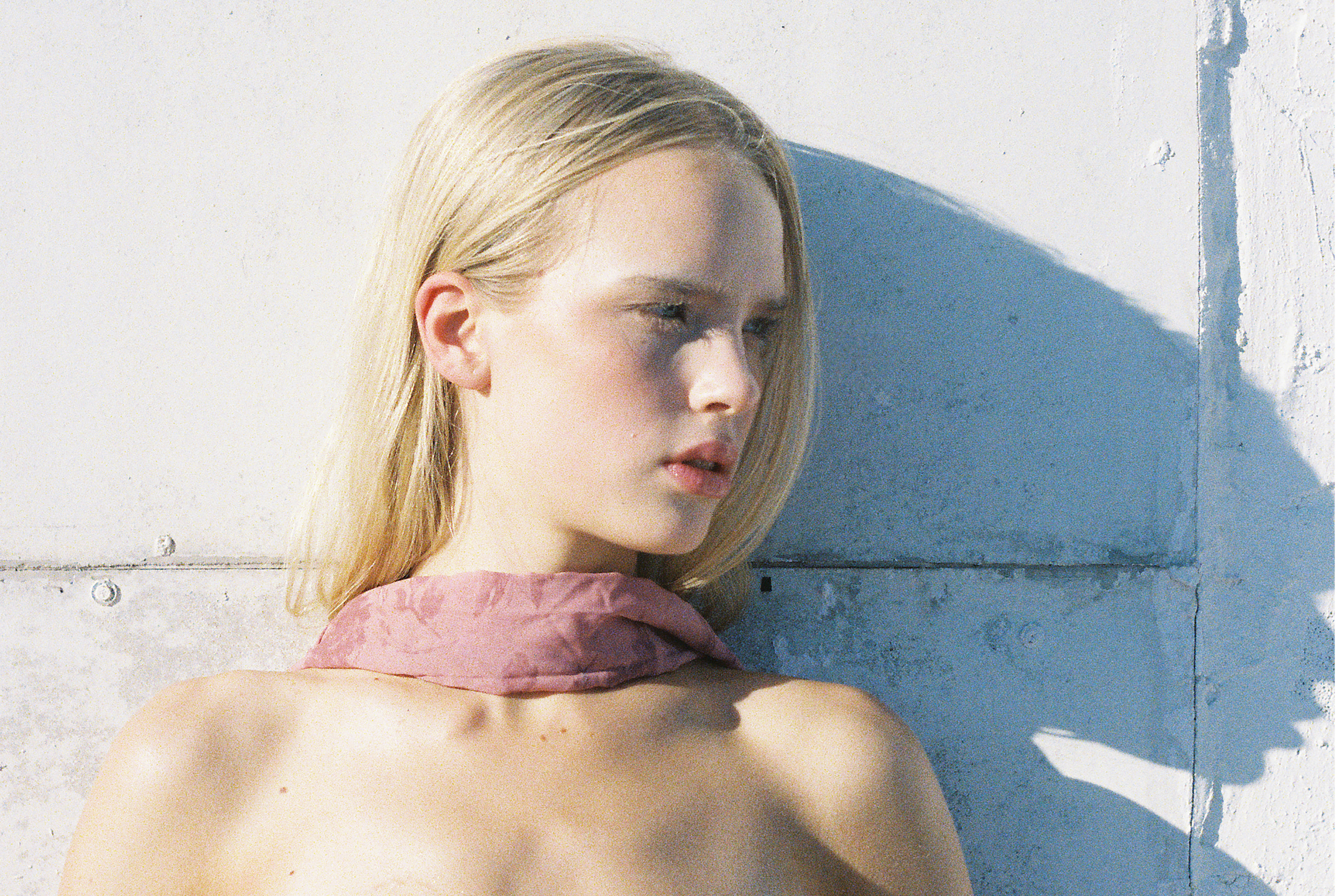 Blonde woman in pink scarf against a textured white wall, deep in thought.