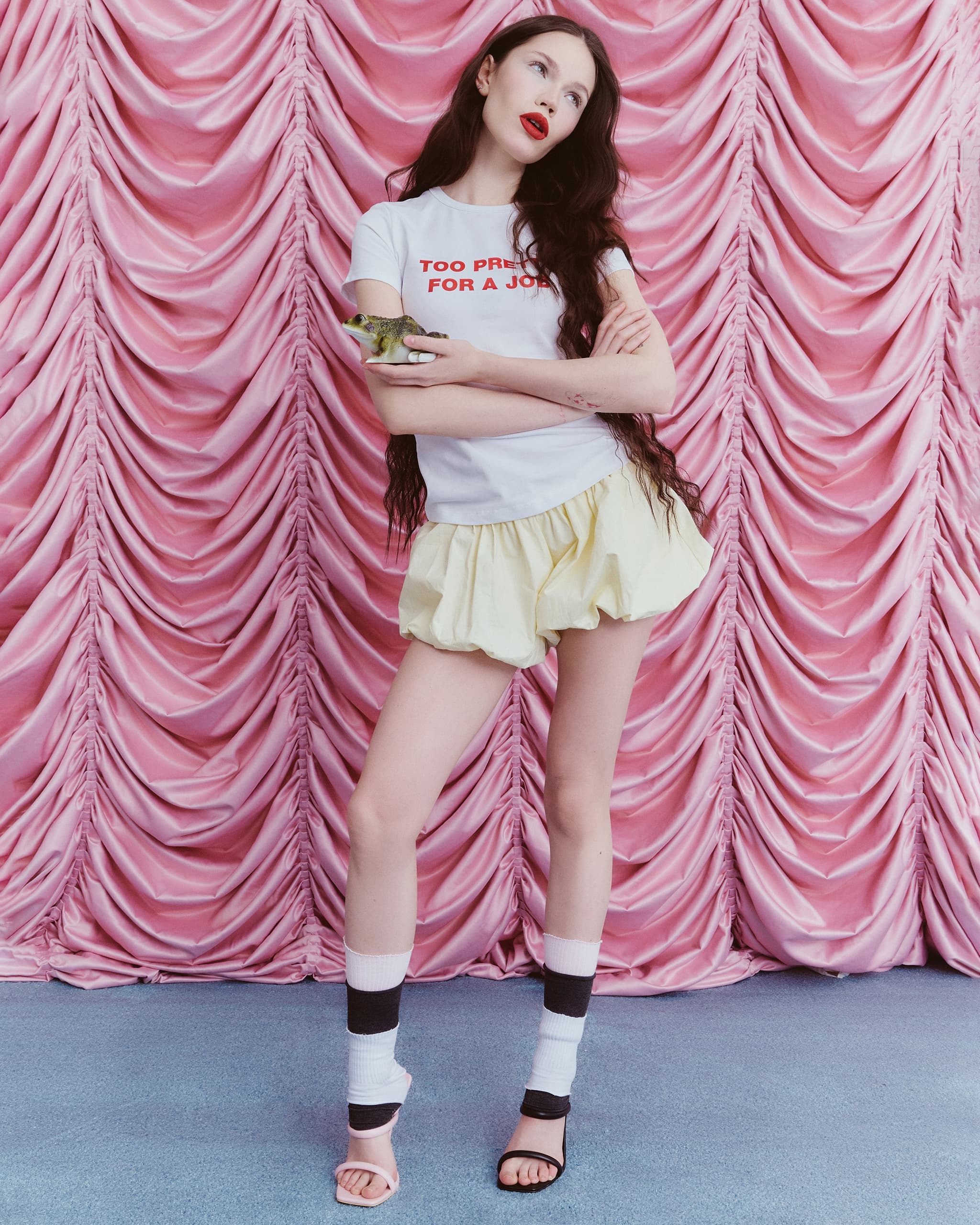 Woman posing with green frog, trendy outfit, pink drapes backdrop.