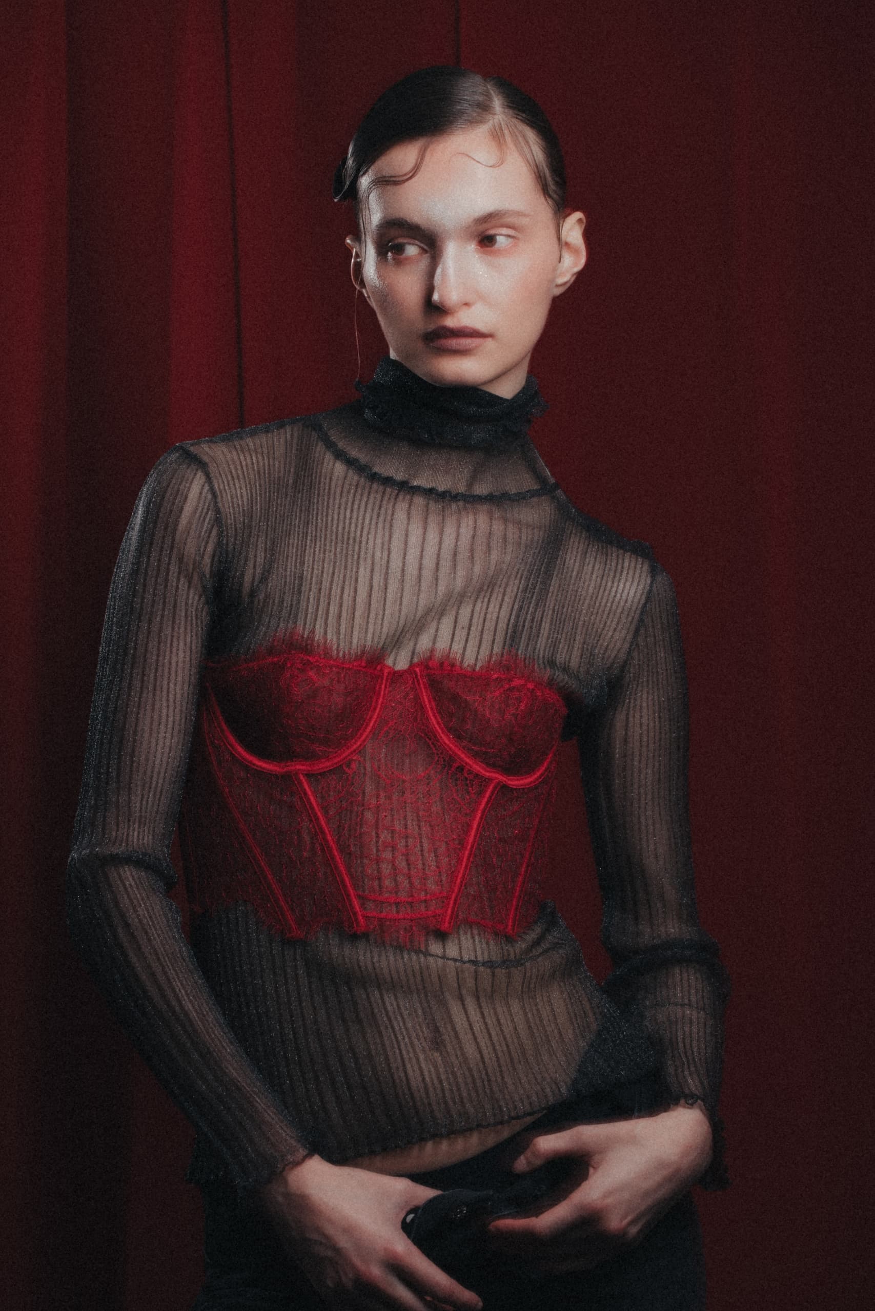 Model in sheer black top with red lace corset, posing against a red curtain backdrop.