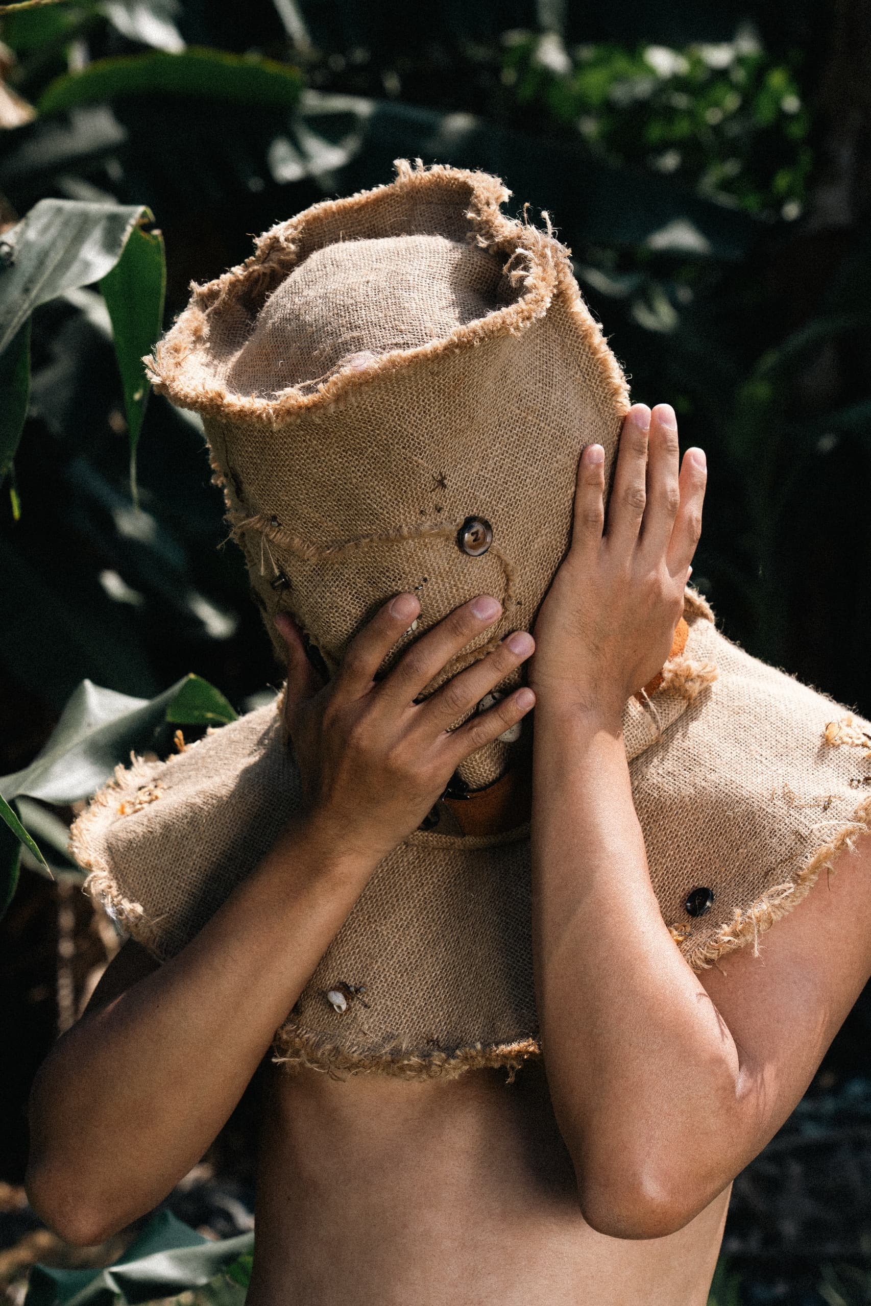 Person in burlap sack costume with hands on face, surrounded by lush greenery.