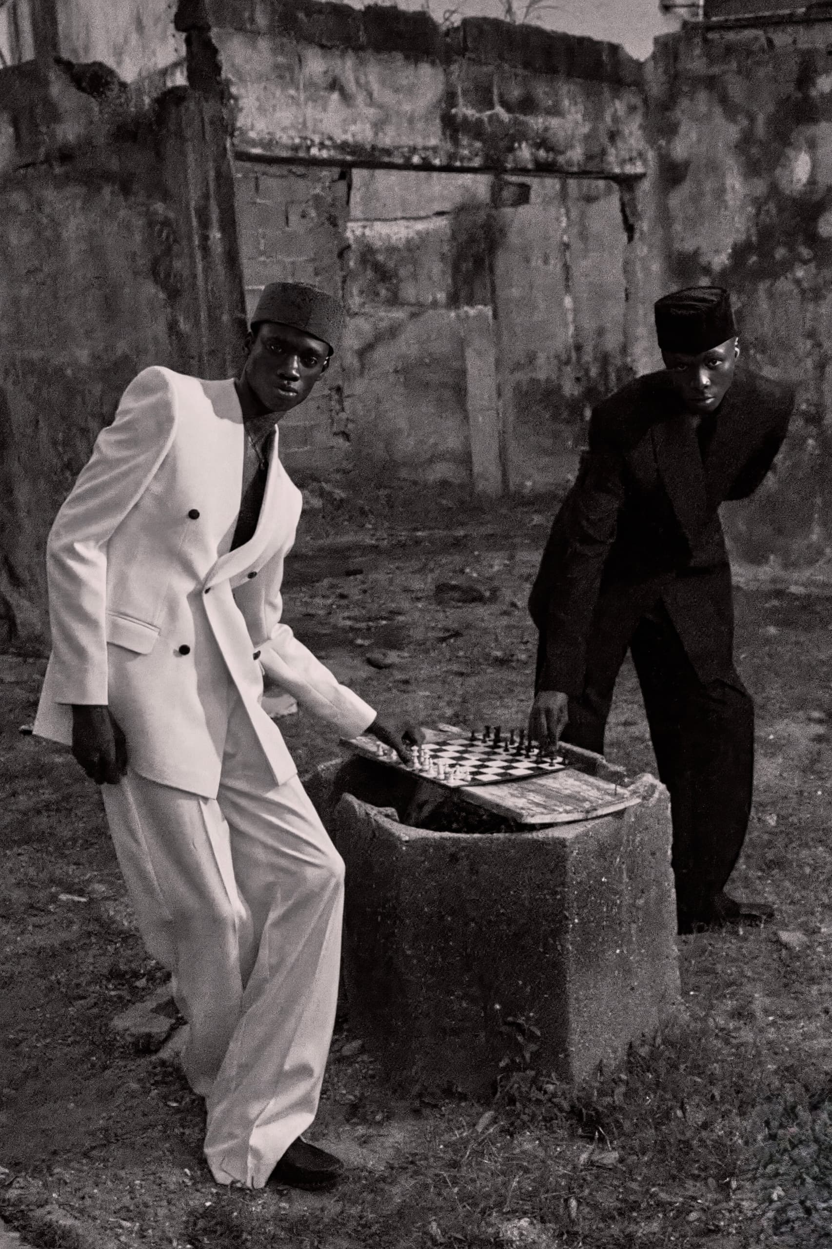 Two men in formal suits playing chess outdoors, with a rustic backdrop.