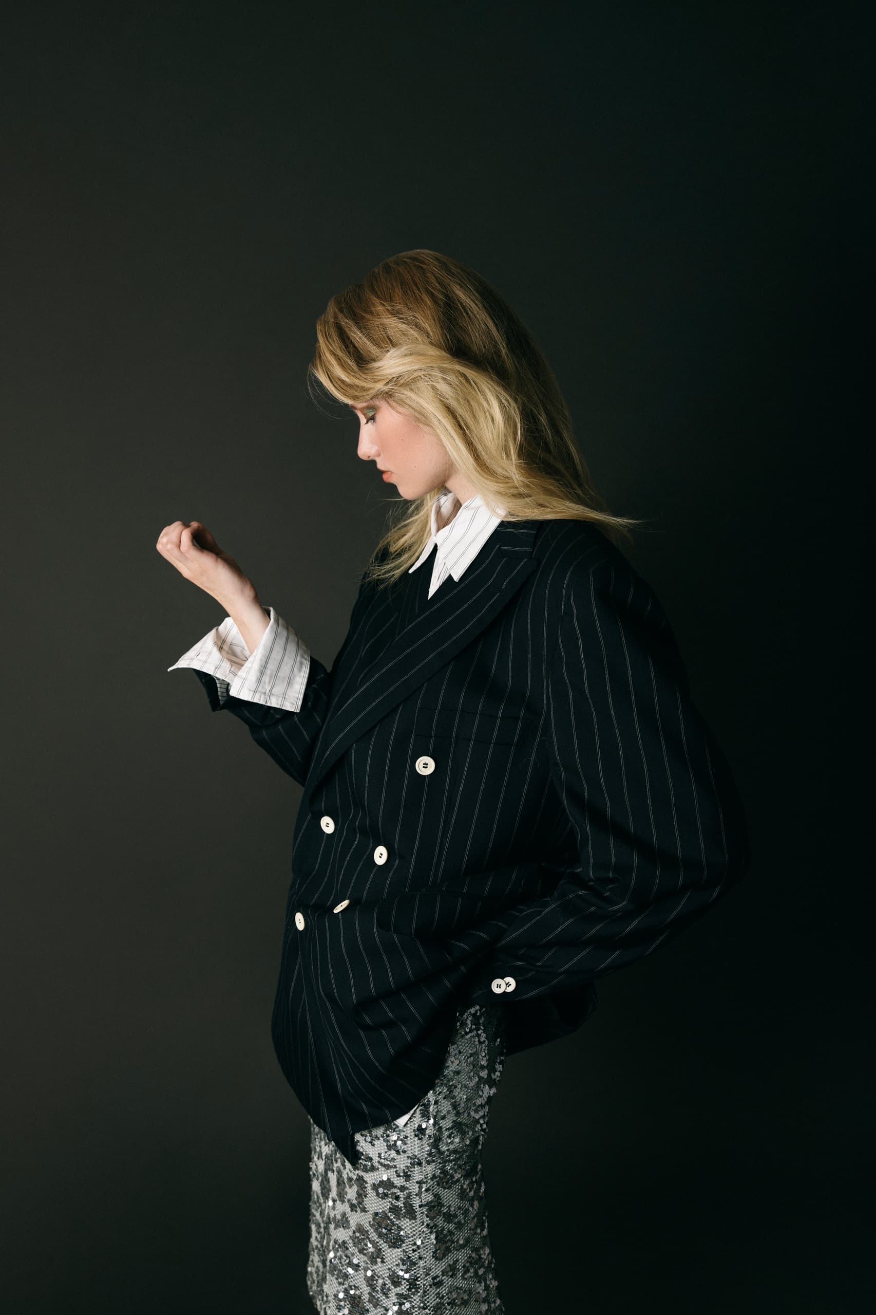 Fashion model in pinstripe blazer and sequin skirt against dark backdrop.