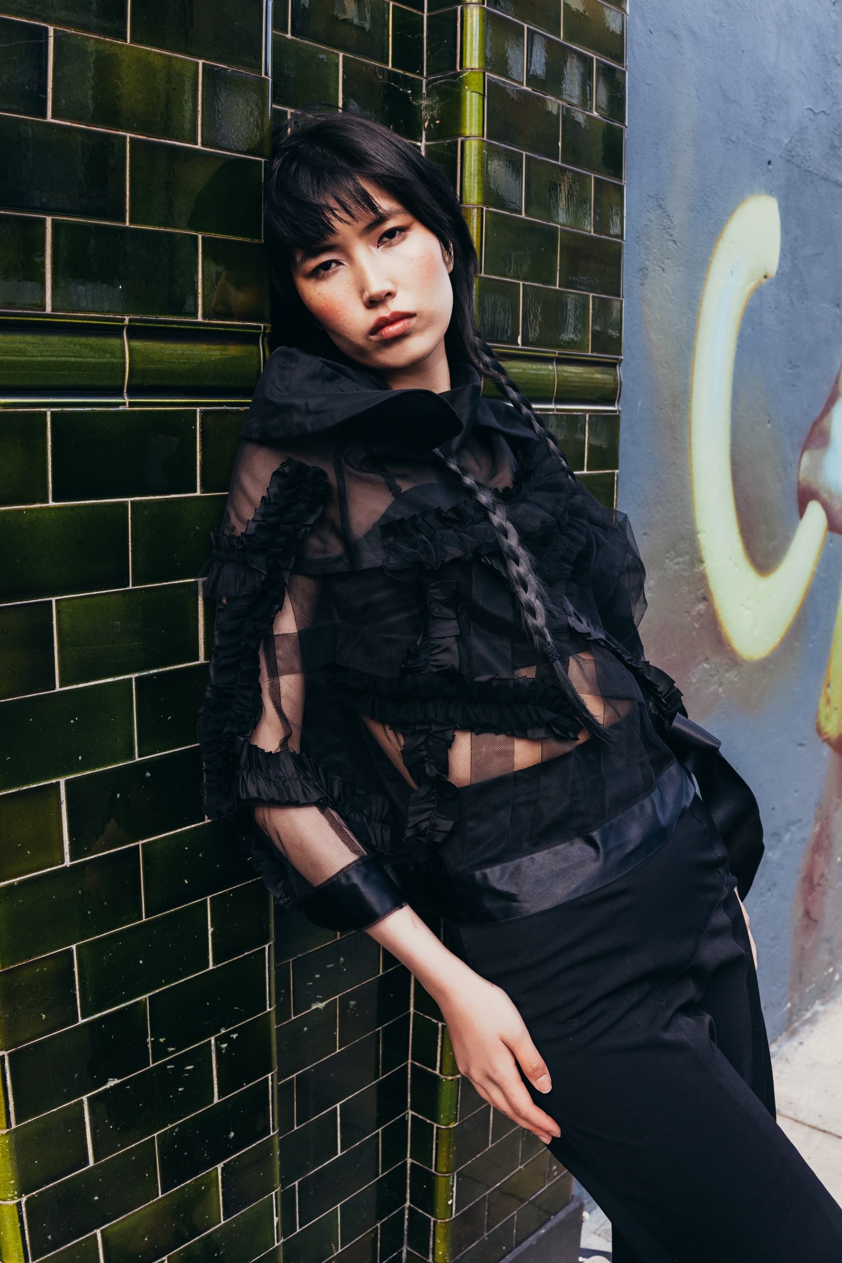Stylish person in black outfit leaning against green-tiled wall with graffiti background.