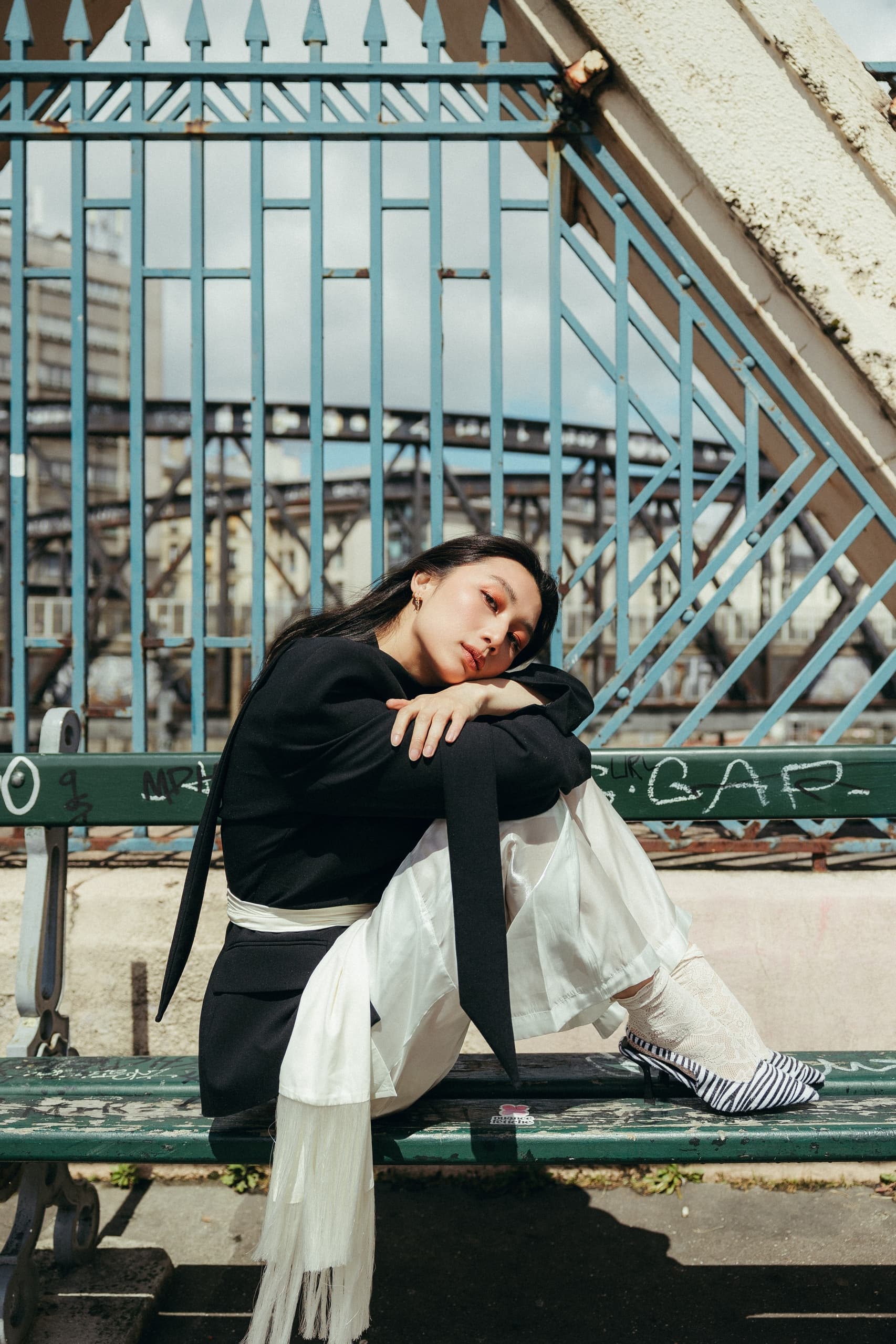 Fashionable woman sitting on a bench in urban setting, wearing stylish monochrome outfit.