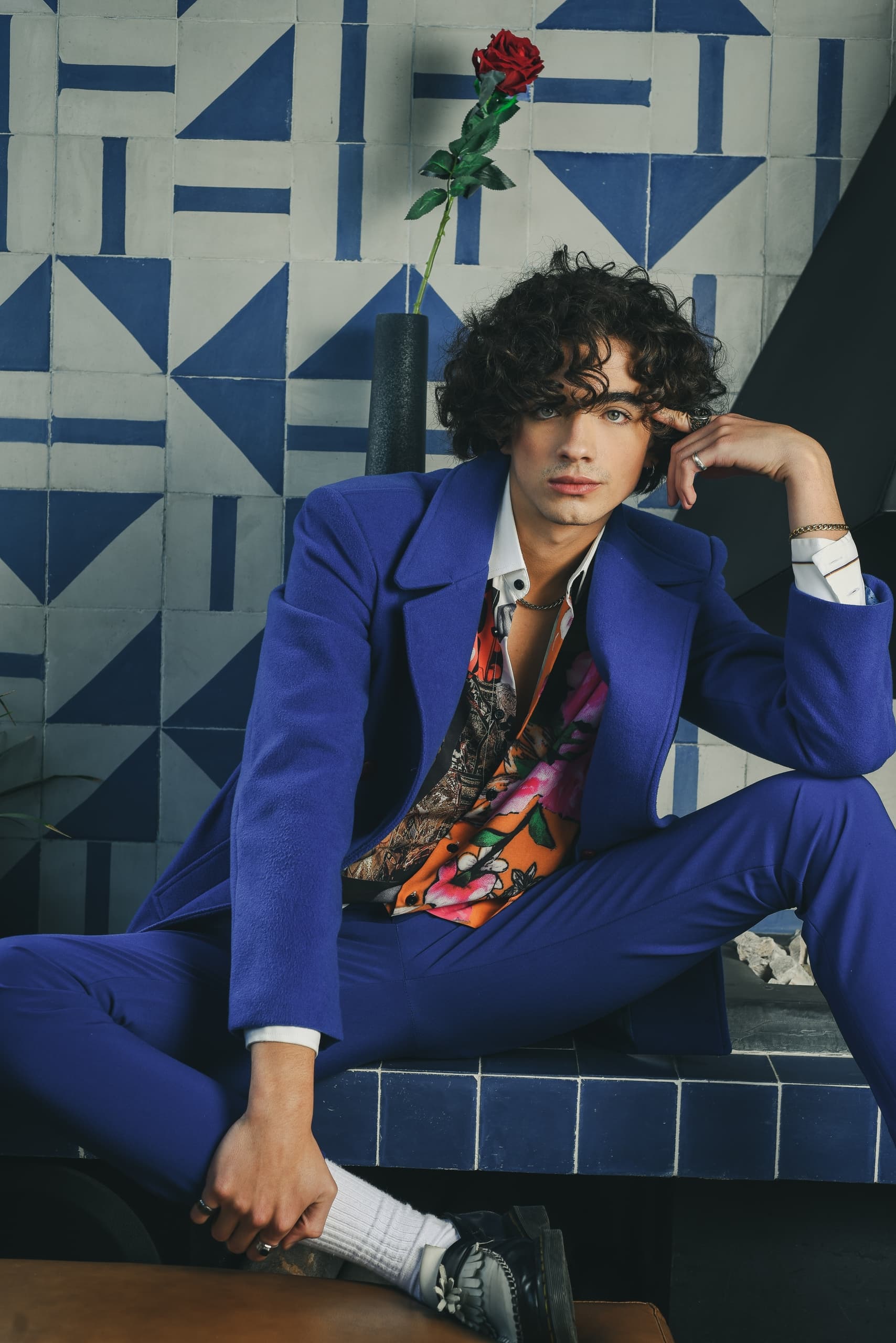 Stylish person in a vibrant outfit with a unique tiled background, holding a pose.