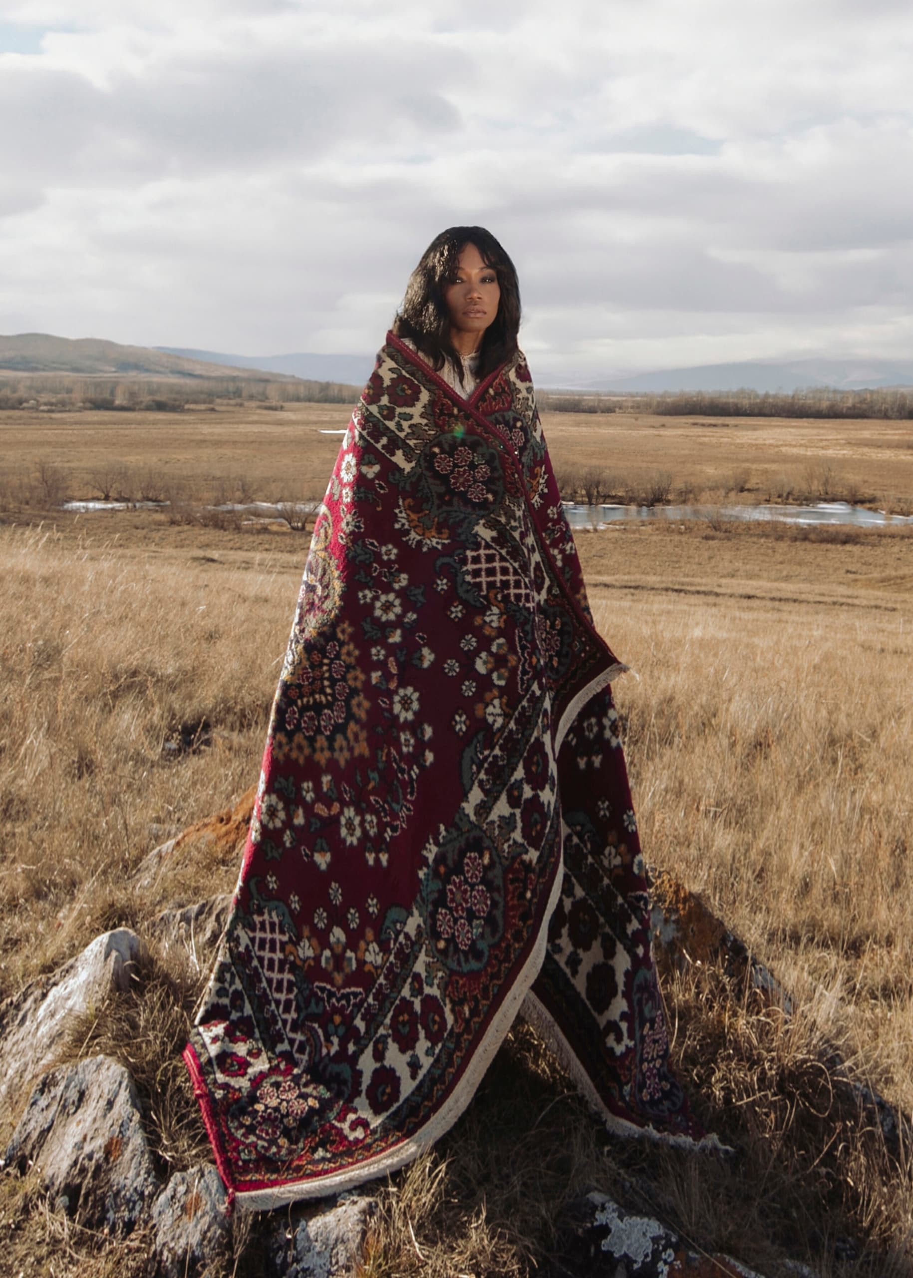 Woman wrapped in a vibrant patterned blanket in a vast, open field.