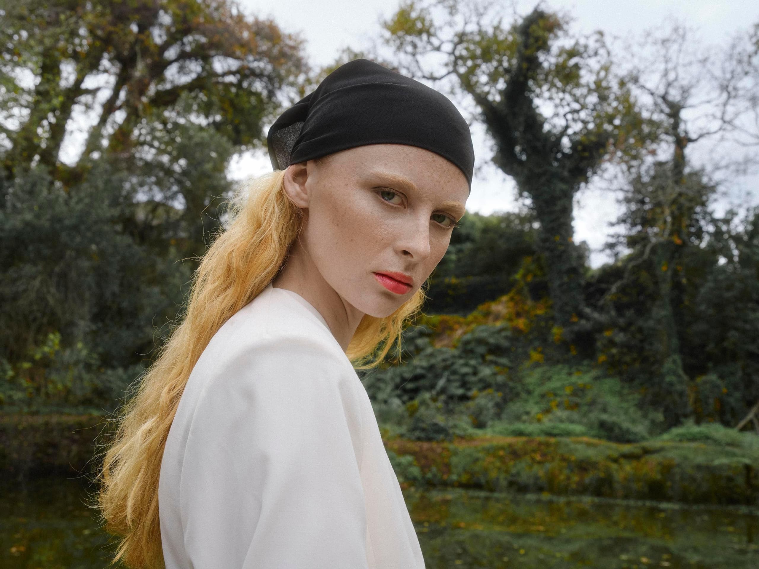 Person in a forest setting wearing a black headscarf and a white outfit.