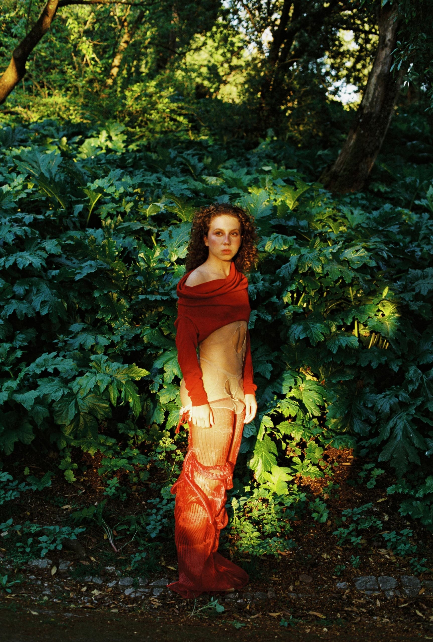 Woman in red outfit stands in lush green forest, sunlight highlighting her face.