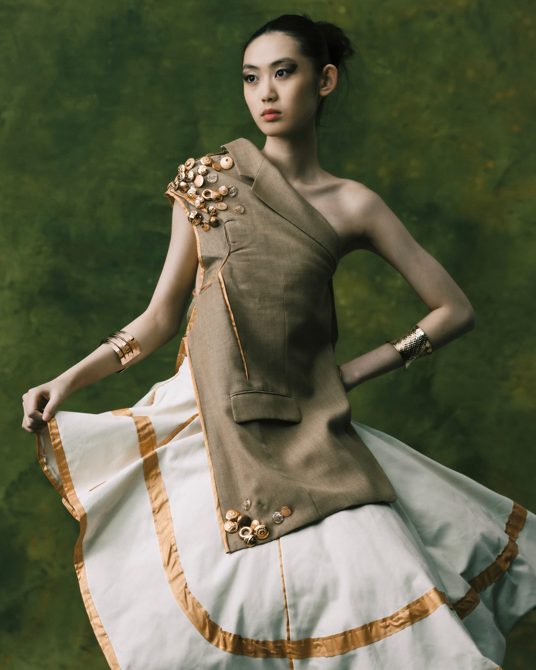 Model in stylish asymmetric dress with button embellishments, posing against green backdrop.