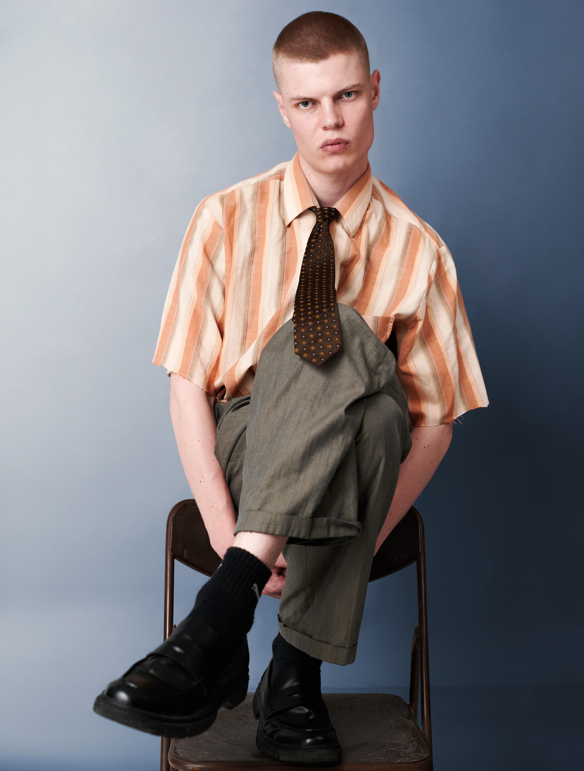 Person in striped shirt and tie sits cross-legged on chair, minimalist fashion style.