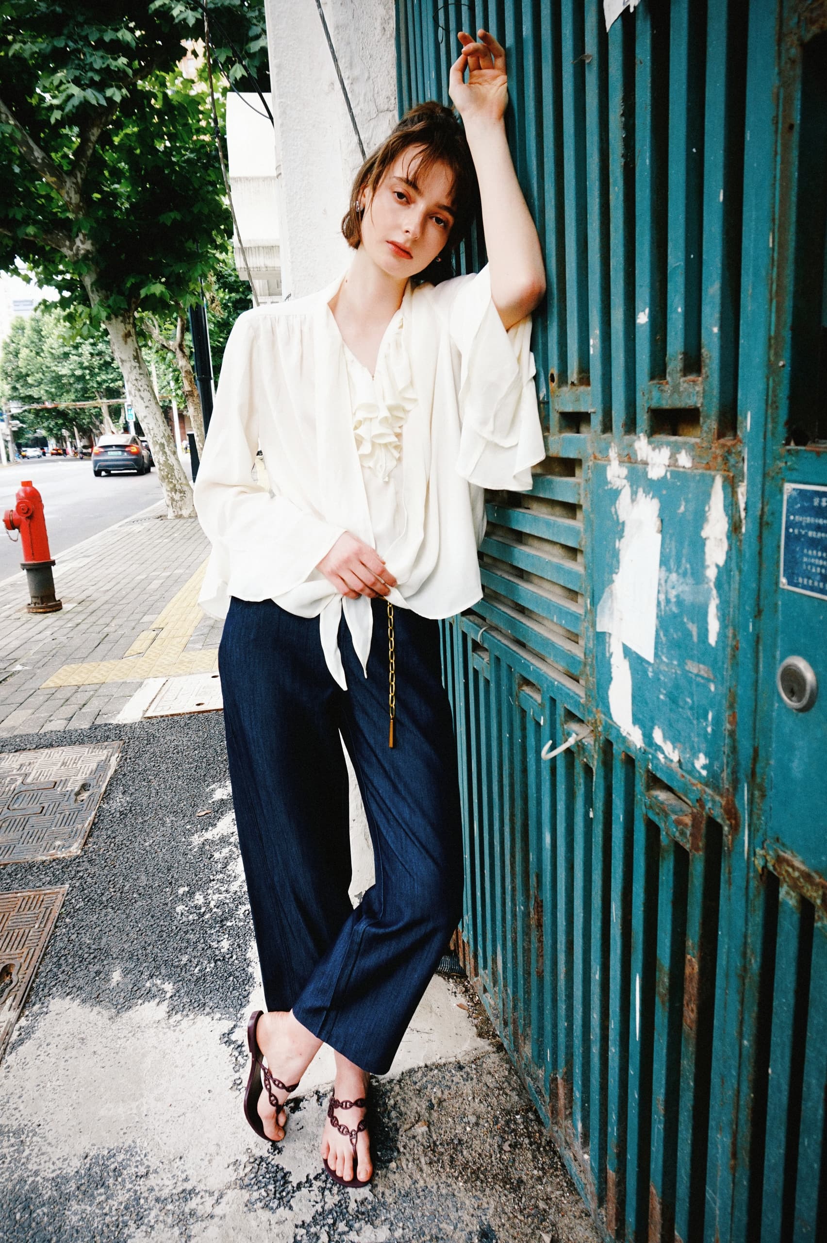 Fashionable woman in white blouse and navy pants leaning against a teal gate.
