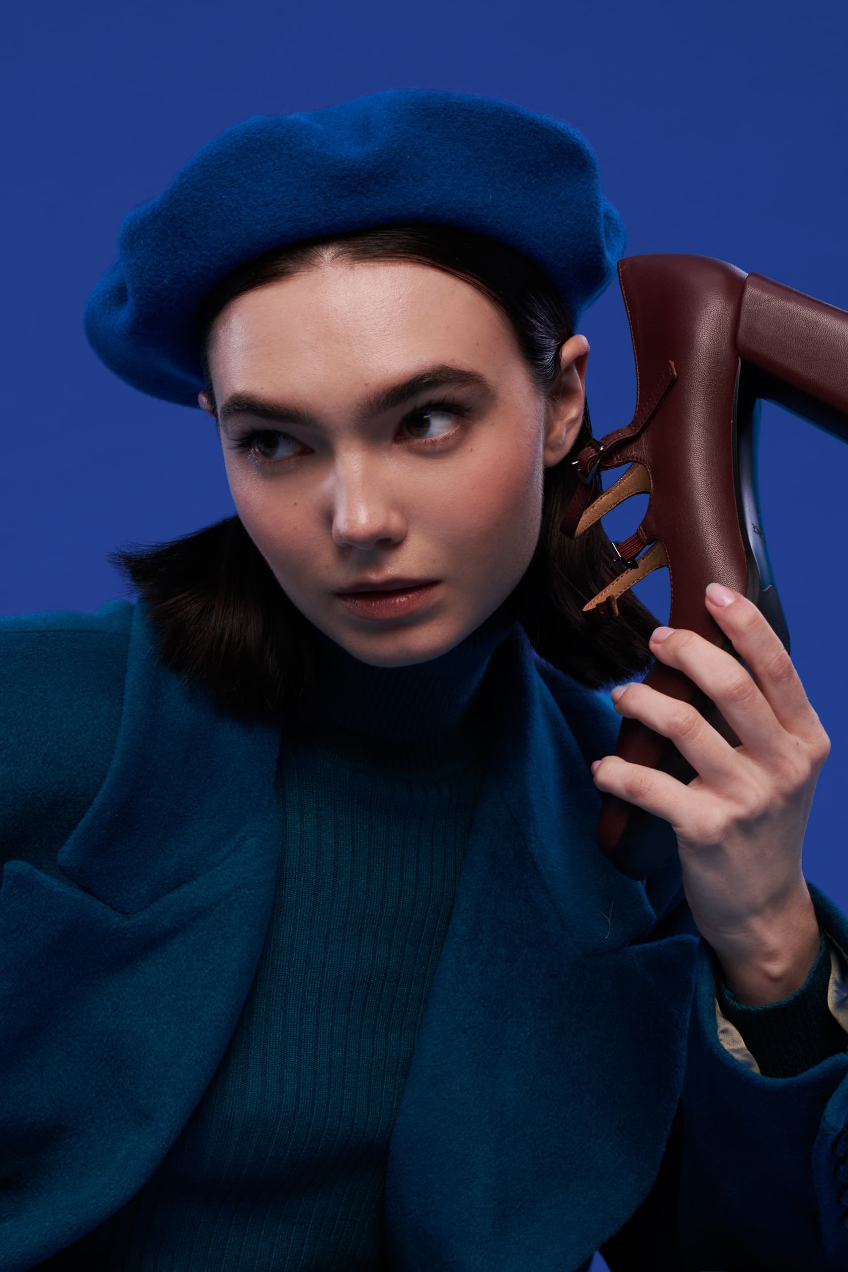 Woman in blue beret uses high heel as a phone against vibrant blue background.