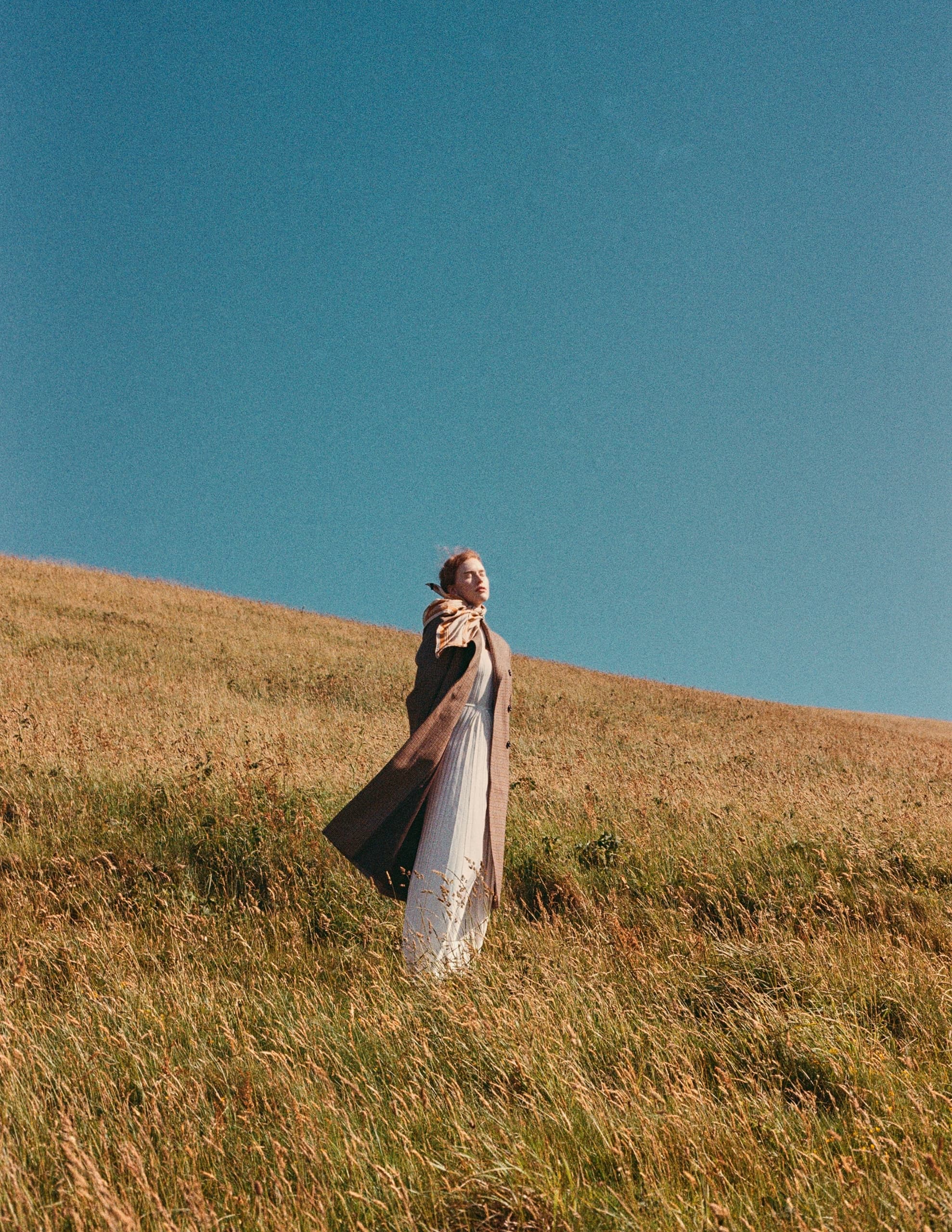 Person in coat standing on grassy hill under clear blue sky.