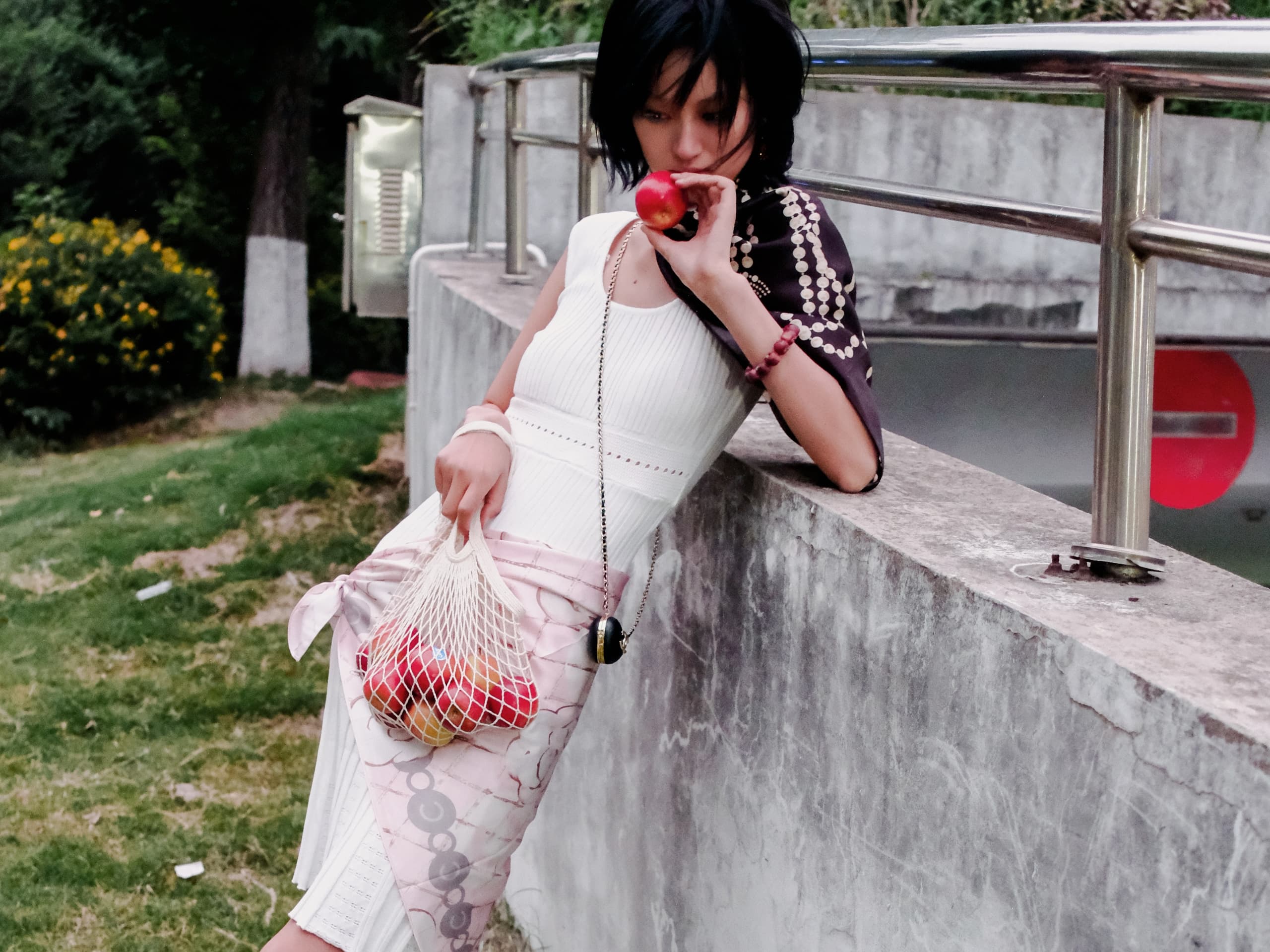 Young woman in stylish attire holding apples, leaning on a concrete wall outdoors.