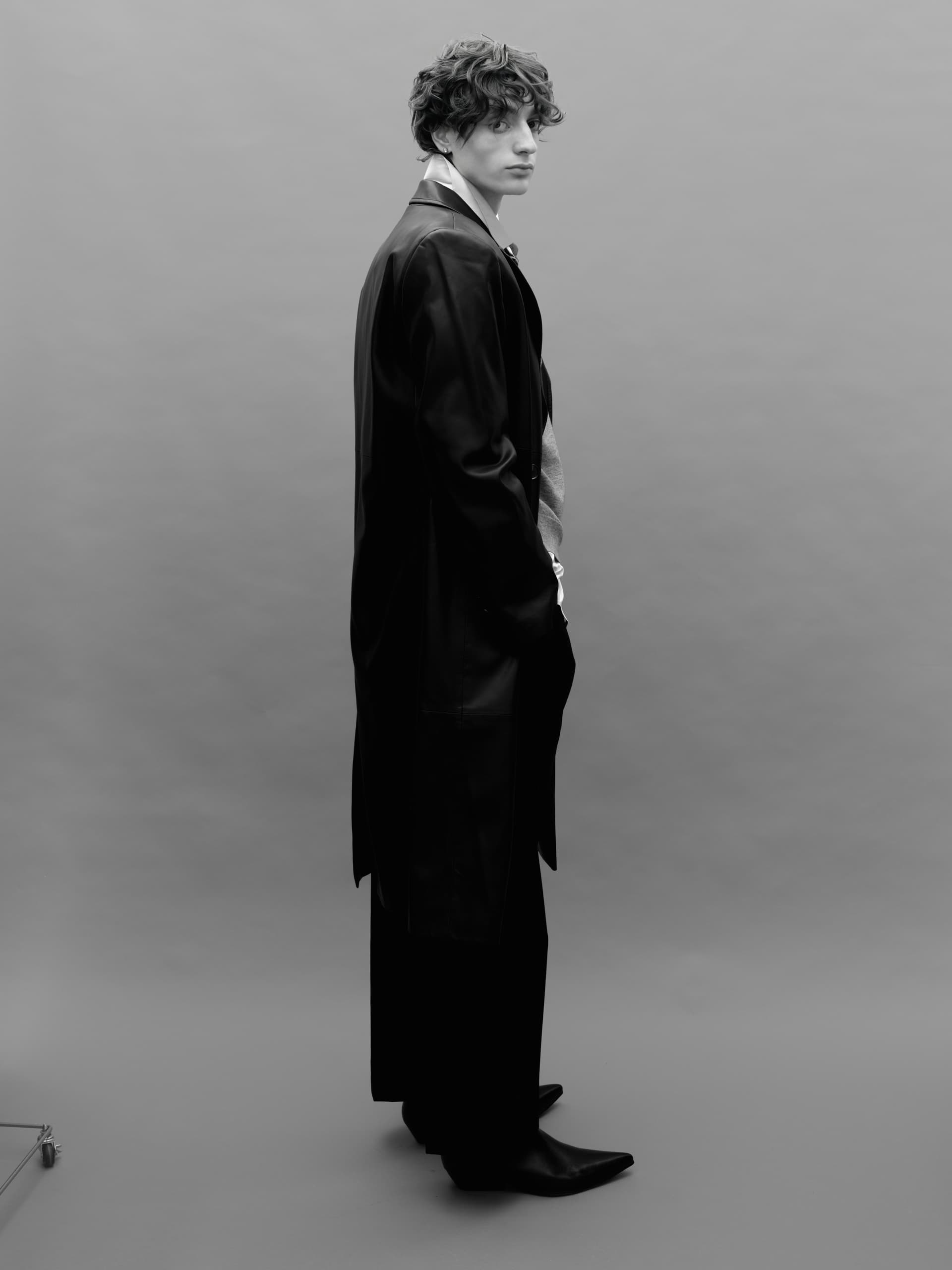 Man in a long black coat poses stylishly against a plain gray background.