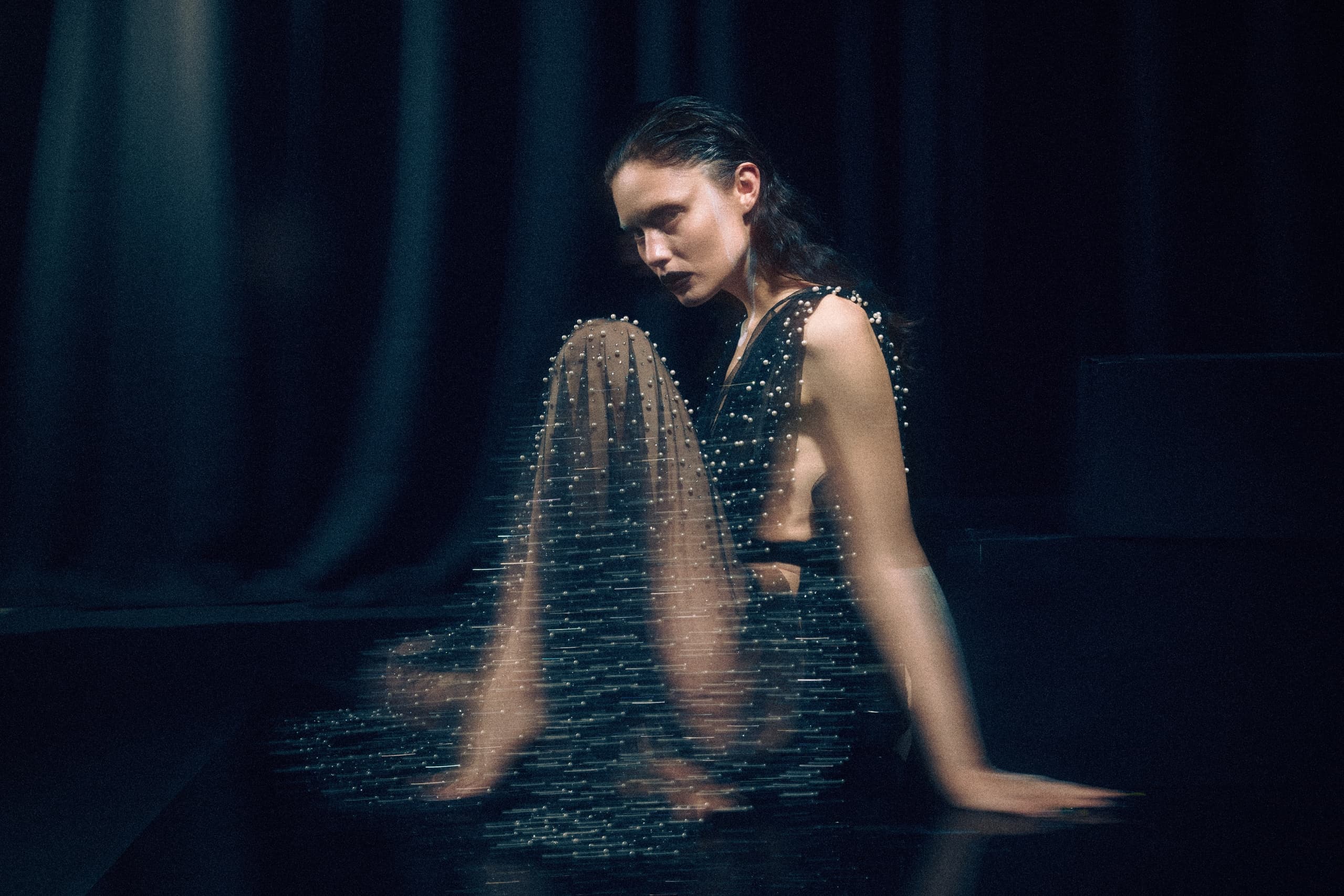 Woman in elegant black dress with pearls, sitting on stage in dim lighting.